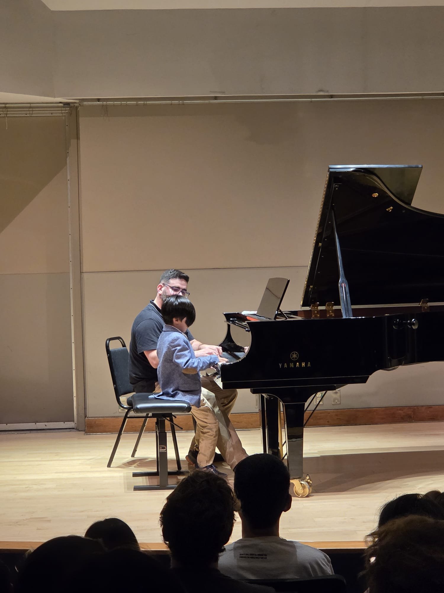 A man and a young boy playing the piano together on stage in front of an audience, with the audience visible in the foreground.