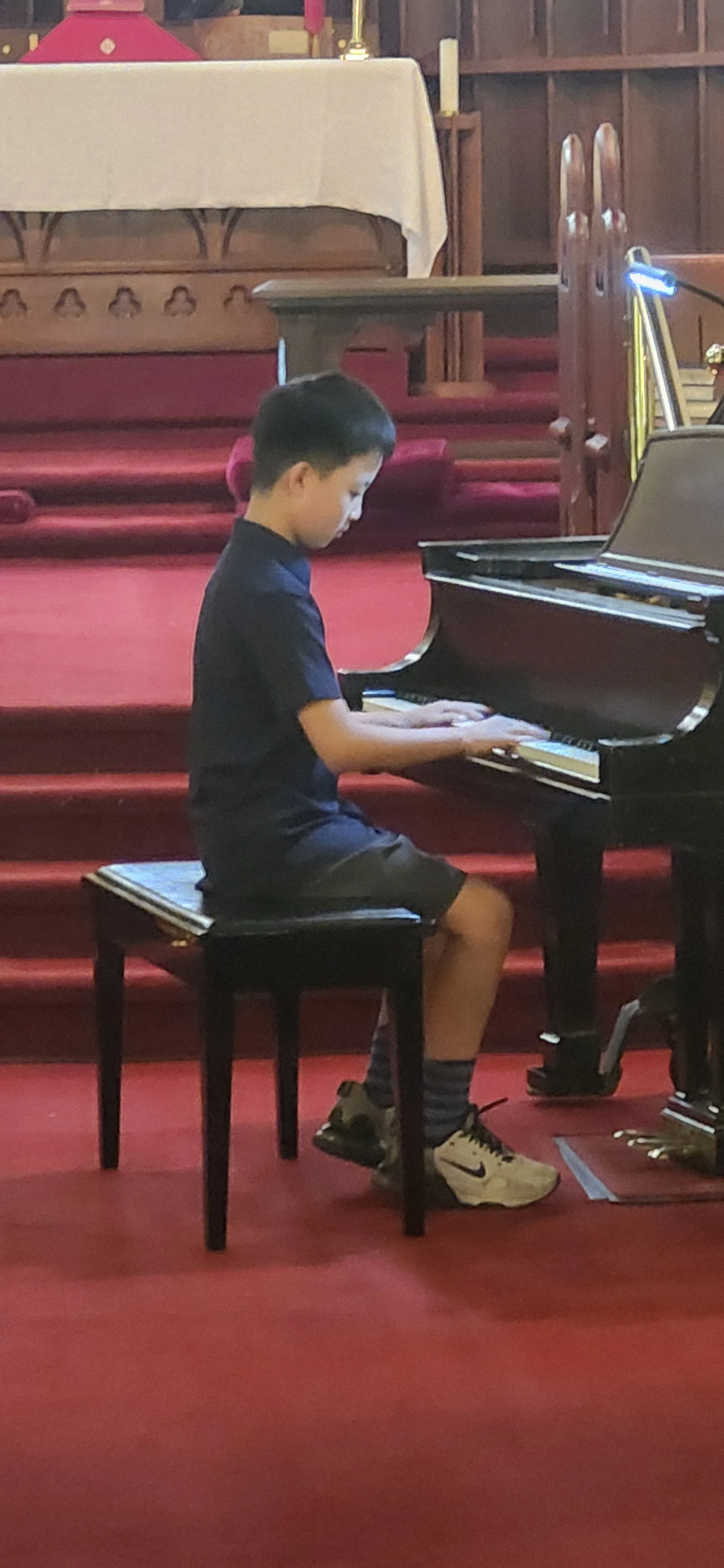 A young boy sitting on a black stool playing a grand piano in a church, with red carpeted stairs and wooden walls in the background.