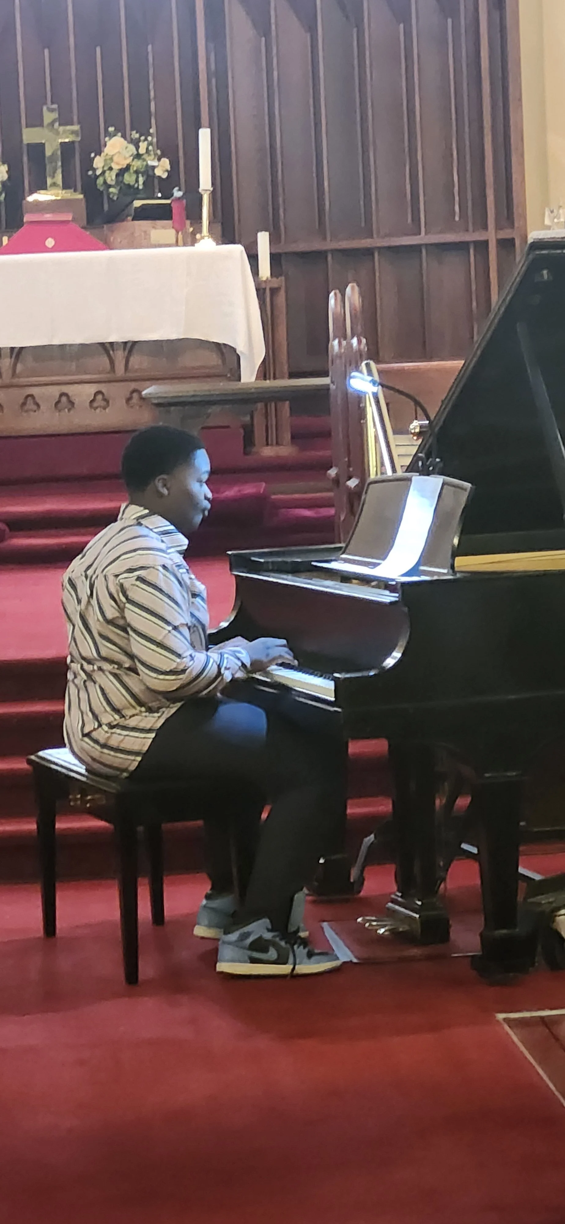 A young person playing a grand piano inside a church with a red carpeted floor, wooden pews, an altar with a cross, flowers, and candles in the background.