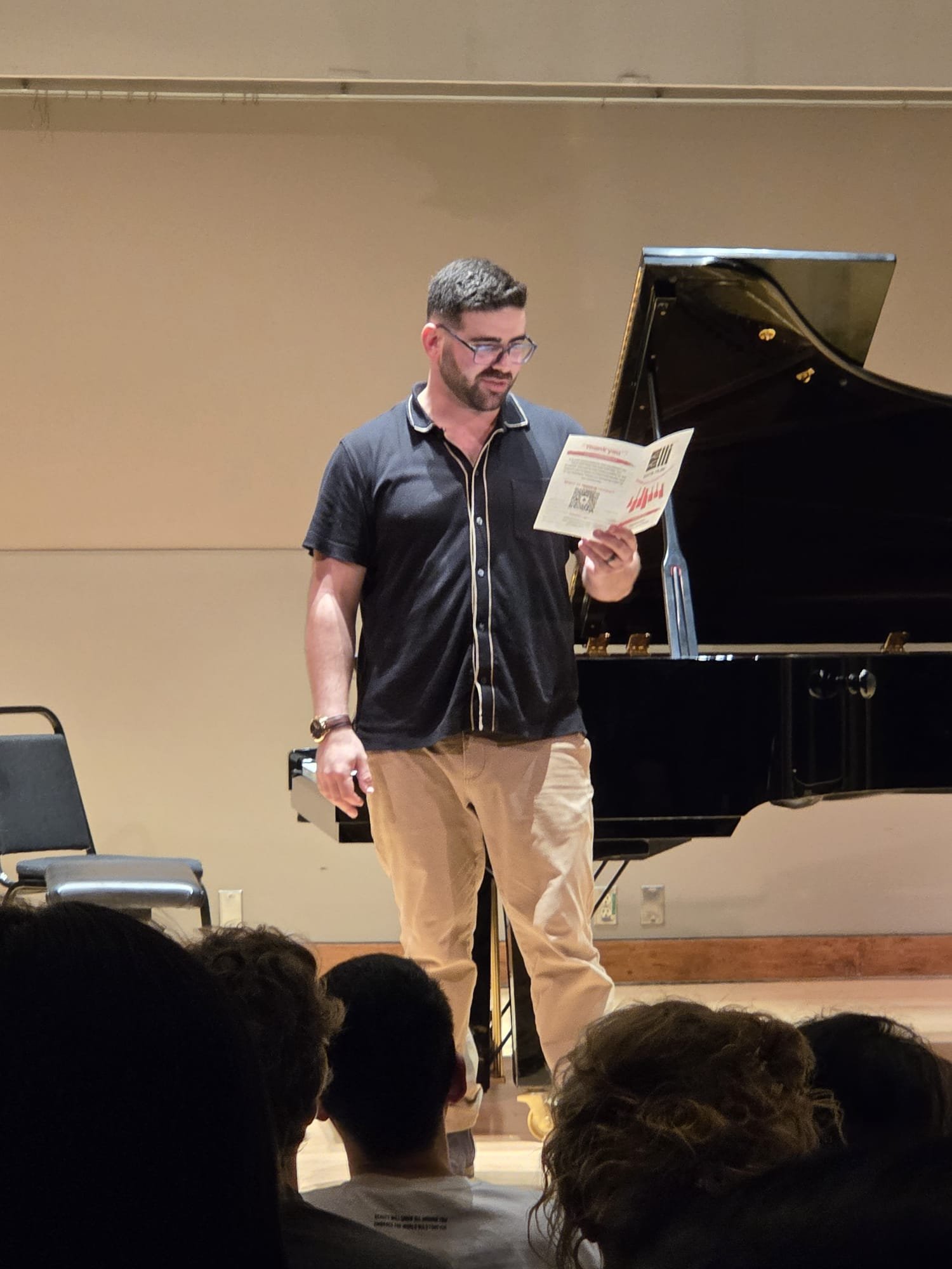 A man with glasses, beard, and short dark hair wearing a black collared shirt and beige pants, reading from a paper in front of a grand piano on stage with an audience in the foreground.