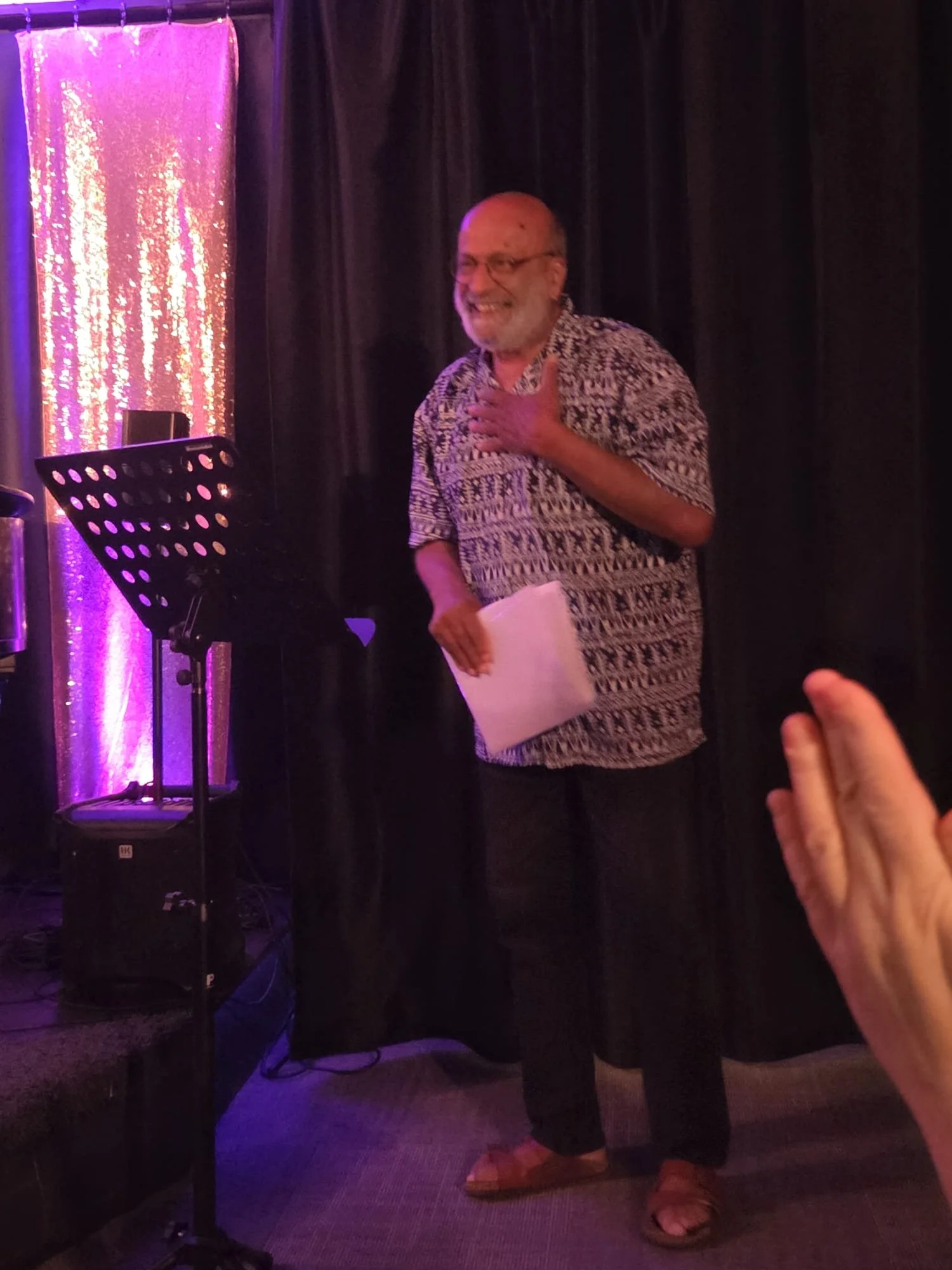 A man with glasses, a beard, and a patterned shirt is smiling and holding a piece of paper, standing in a dimly lit room with a black curtain and a shimmering pink backdrop. There is a music stand and other equipment nearby, and a hand is reaching to
