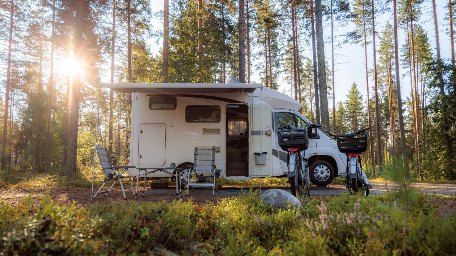 A white camper van parked in a forest with sunlight filtering through tall trees, surrounded by outdoor chairs, a table, and bicycles.