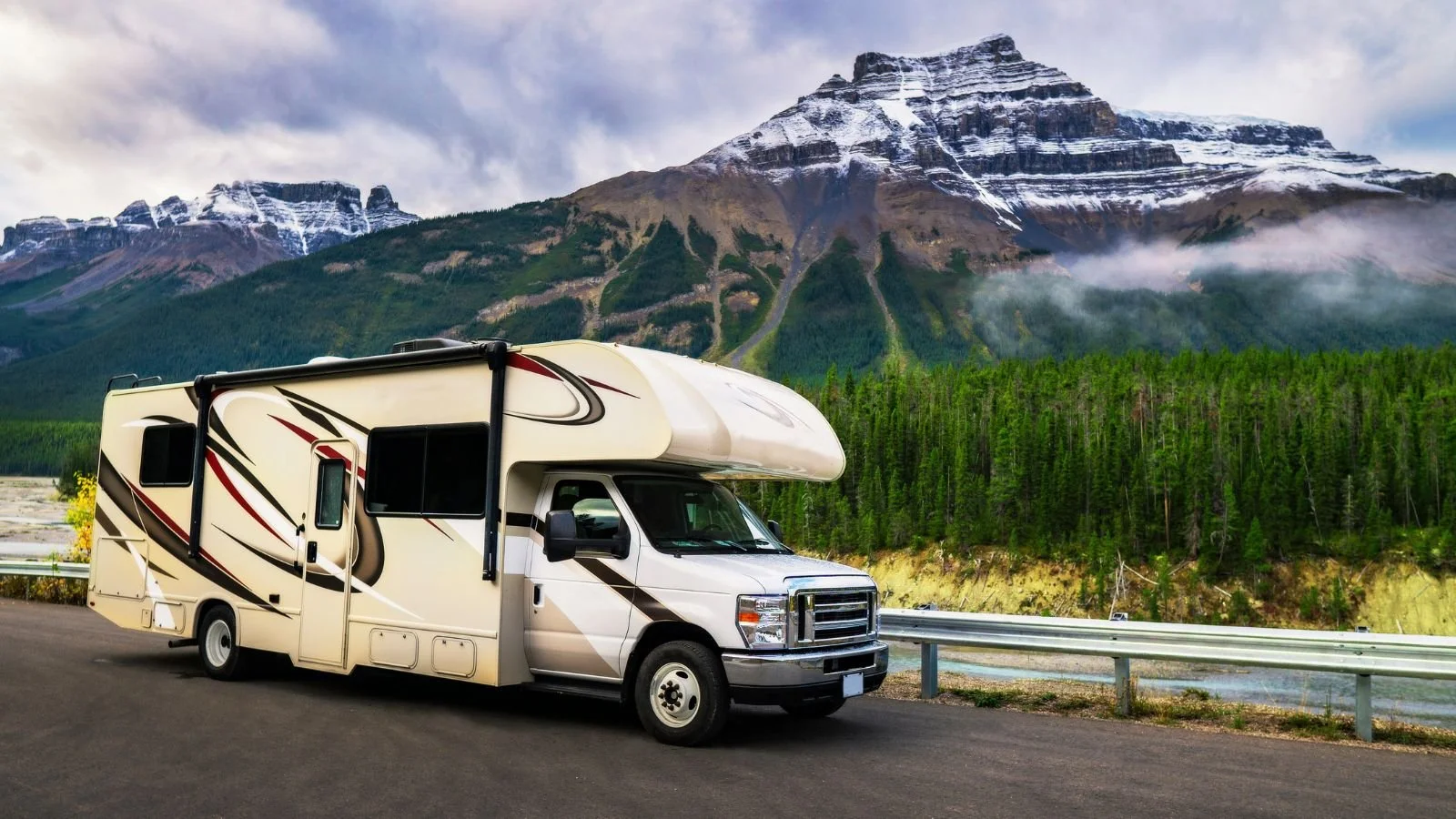 A white motorhome parked on a roadside near a forest with mountainous terrain and snow-capped peaks in the background.