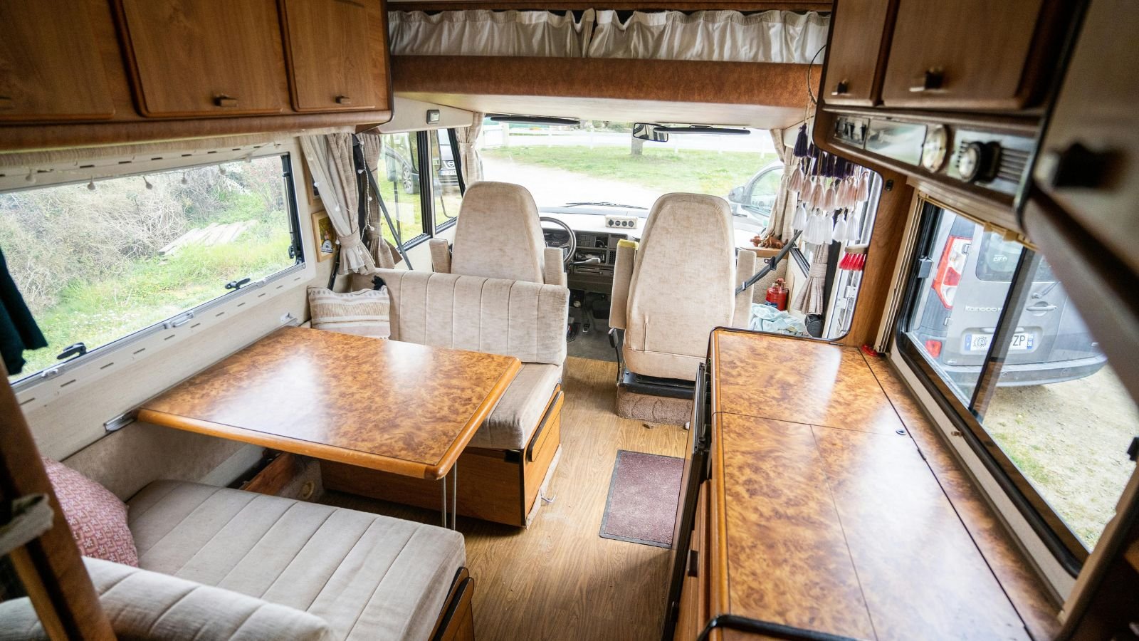Interior of a vintage camper van with wood-paneled cabinets, a dining table, and seating area near large windows showing an outdoor view of grass and parked cars.