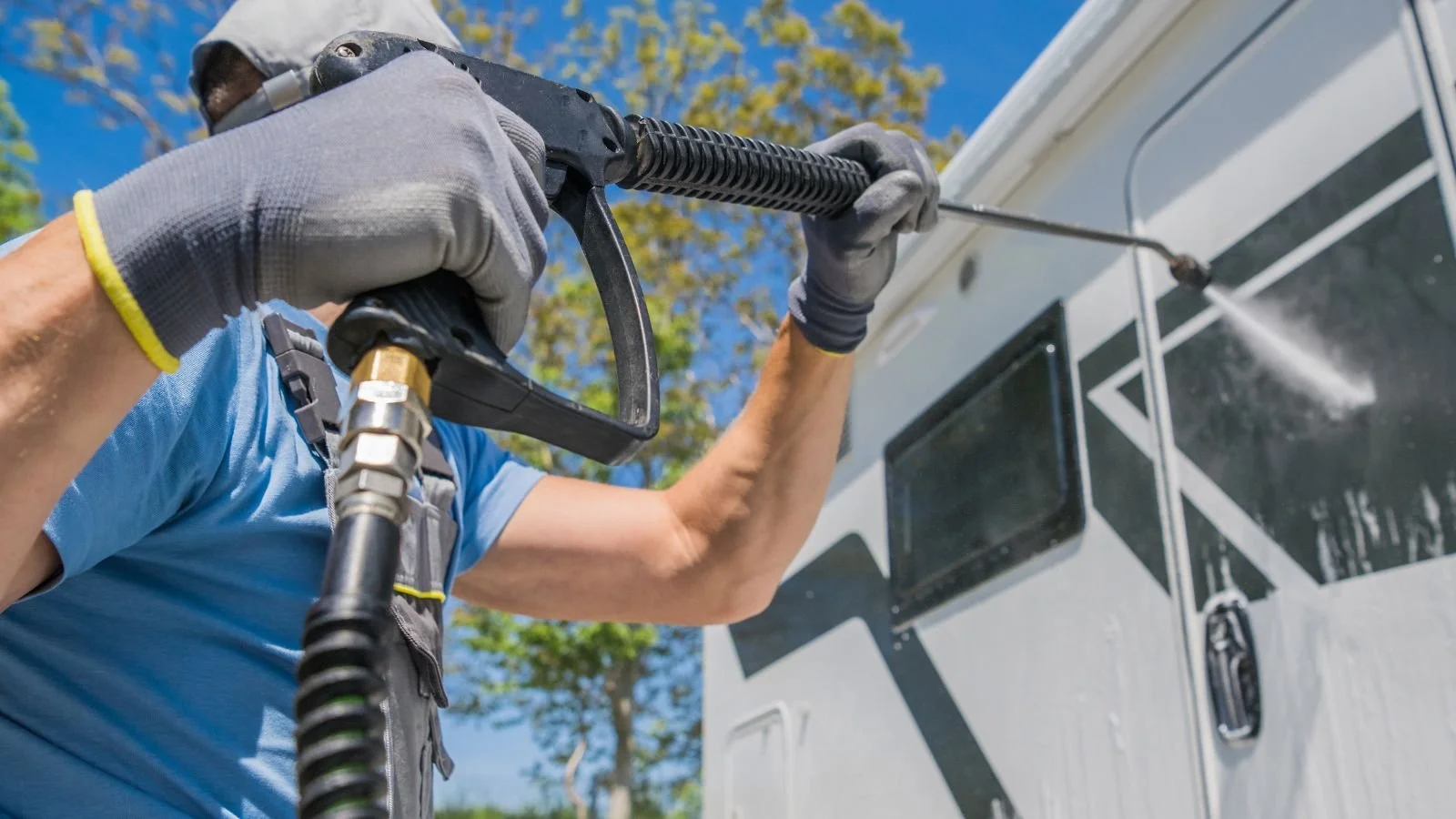Person using a pressure washer to clean the exterior of a camper or RV in an outdoor setting.