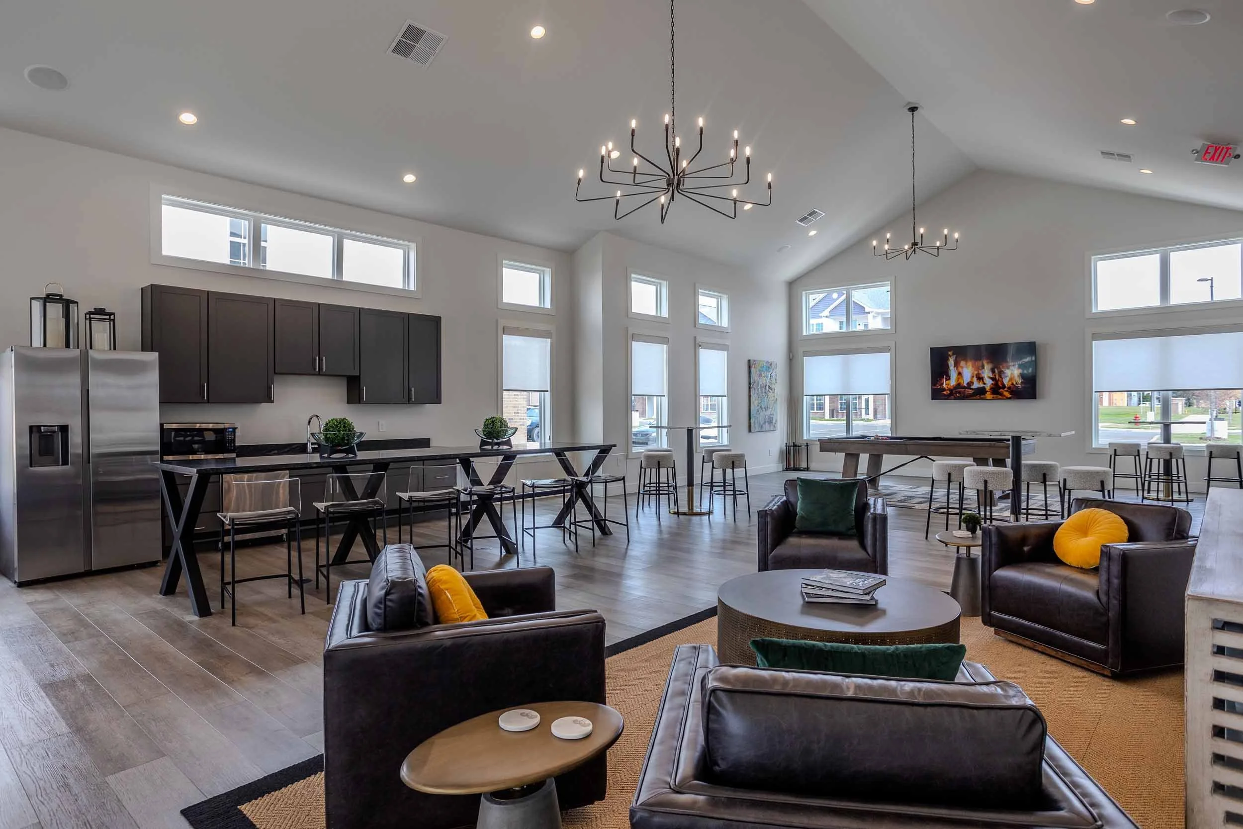 Modern open-concept living space with high ceilings, large windows, two black chandeliers, a kitchen area with dark cabinets, stainless steel refrigerator, black countertop, and bar stools, and a living area with leather chairs, a wall-mounted TV, and decorative pillows.