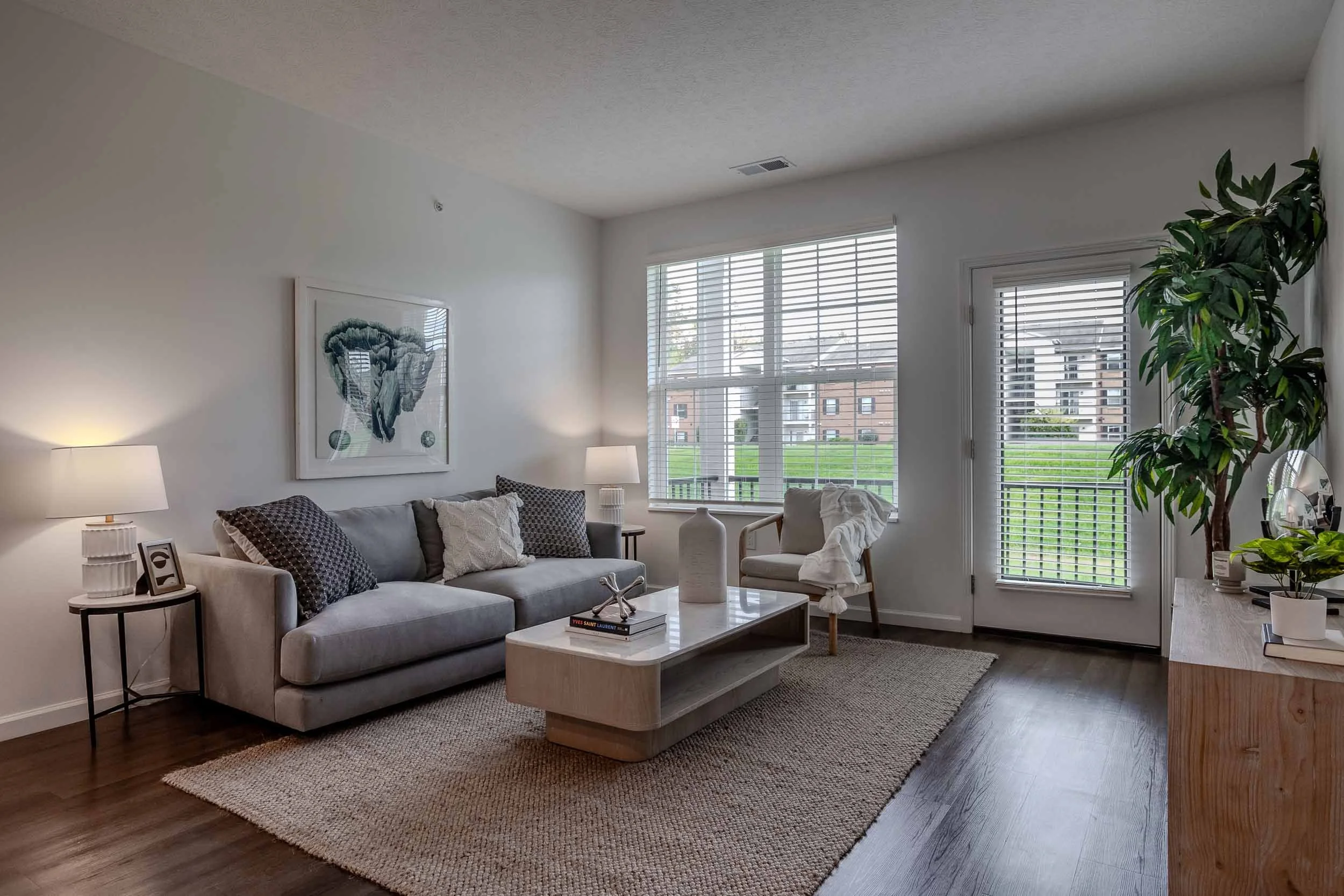 Living room with a beige sofa with patterned pillows, a wooden coffee table, a side table with a photo frame and lamp, white walls with art, large windows with blinds, a potted plant, a white armchair, and a textured area rug, bright natural light outside.
