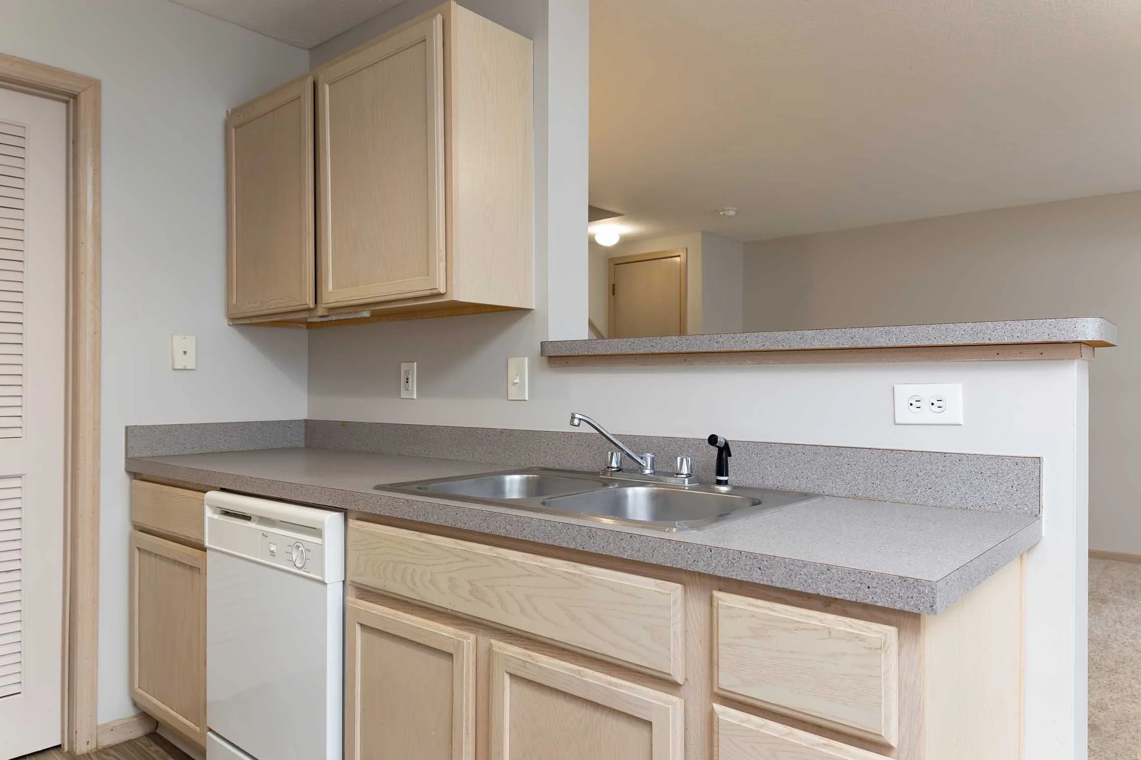 A kitchen with wooden cabinets, a double sink, a dishwasher, and electrical outlets on a speckled gray countertop, with a view into an adjacent room.