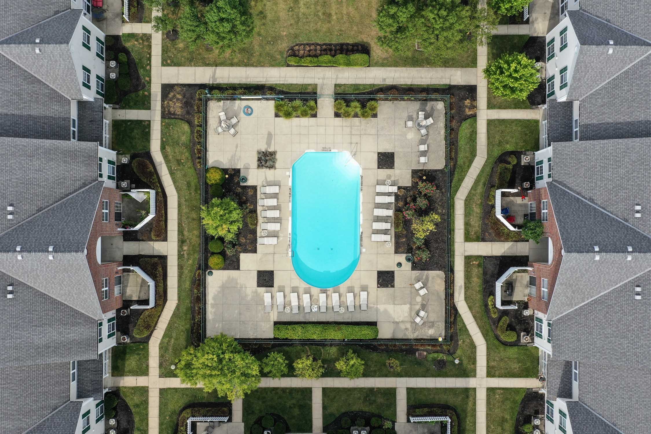 Aerial view of an apartment complex courtyard featuring a swimming pool in the center, surrounded by lounge chairs and lush greenery, with sidewalks and multi-story residential buildings.