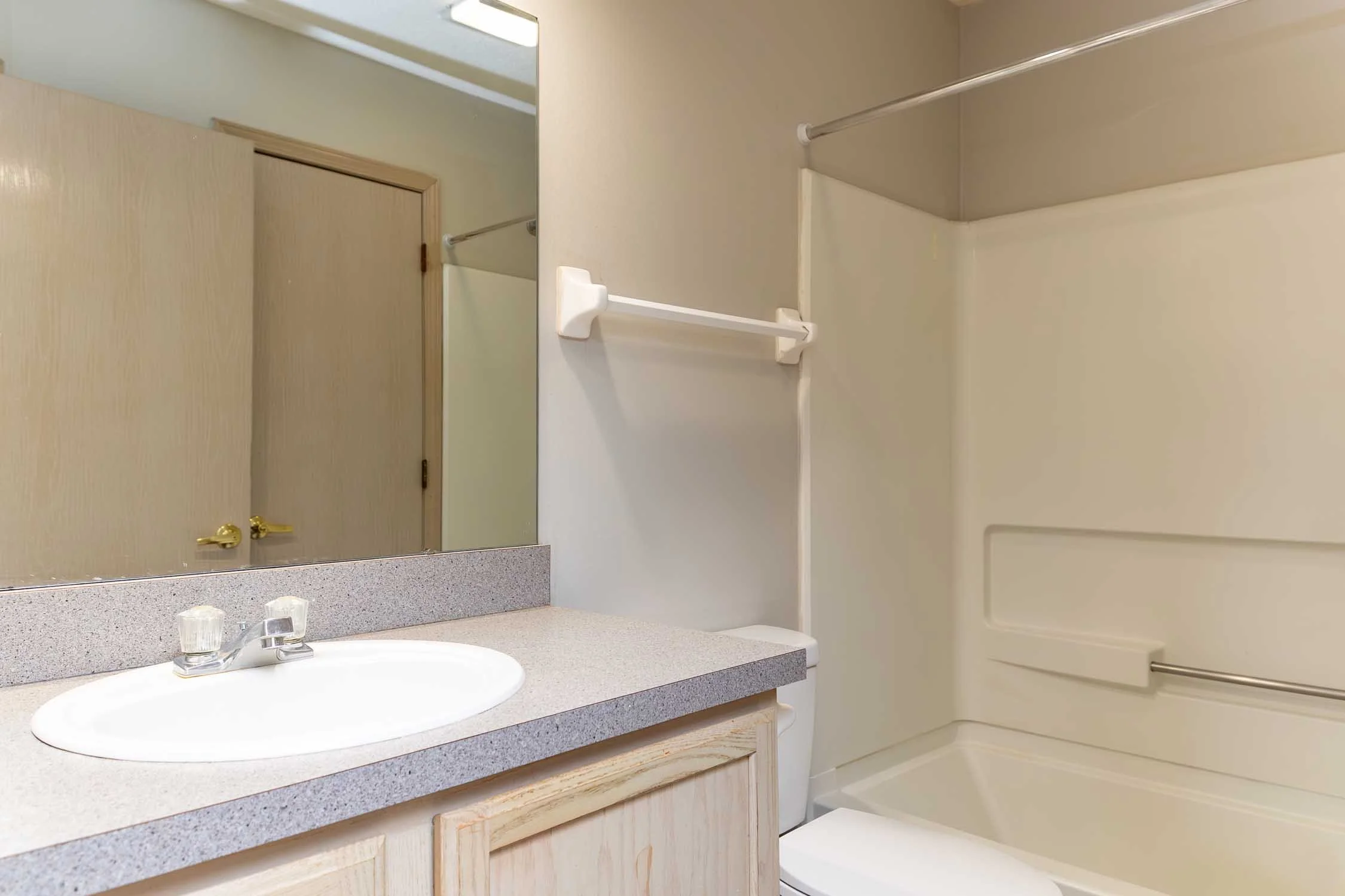 Bathroom with beige walls, a mirror above a speckled countertop with an oval sink, two clear plastic cups, a faucet, and wood cabinets below. To the right, there is a shower or bathtub with a beige surround and a metal grab bar.