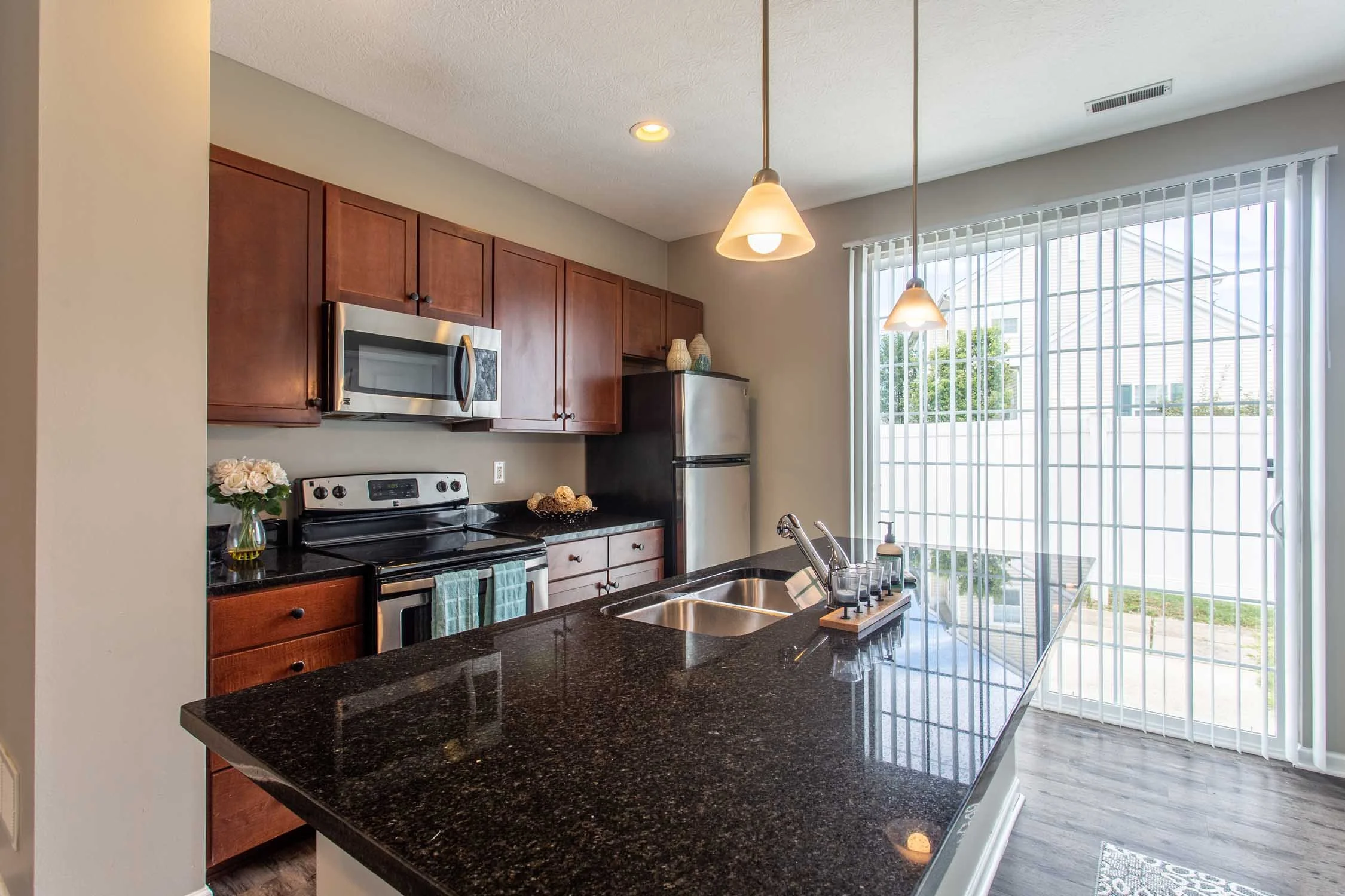 Kitchen with dark granite countertops, wooden cabinets, stainless steel microwave and refrigerator, a window with vertical blinds, and pendant lights.