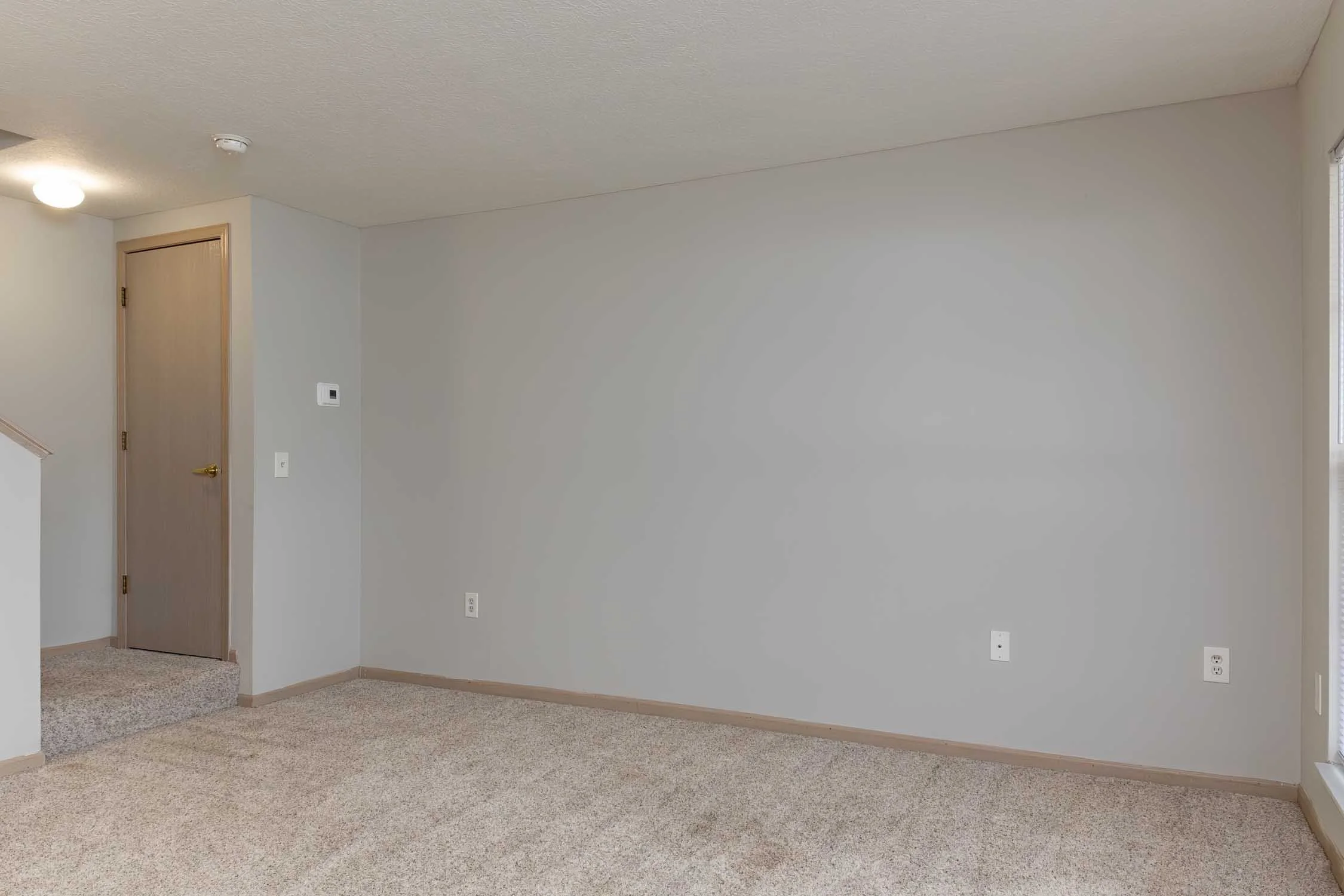 Empty room with beige carpet, light gray walls, and a wooden door. There are multiple electrical outlets and a thermostat on the wall. Part of a staircase is visible on the left.