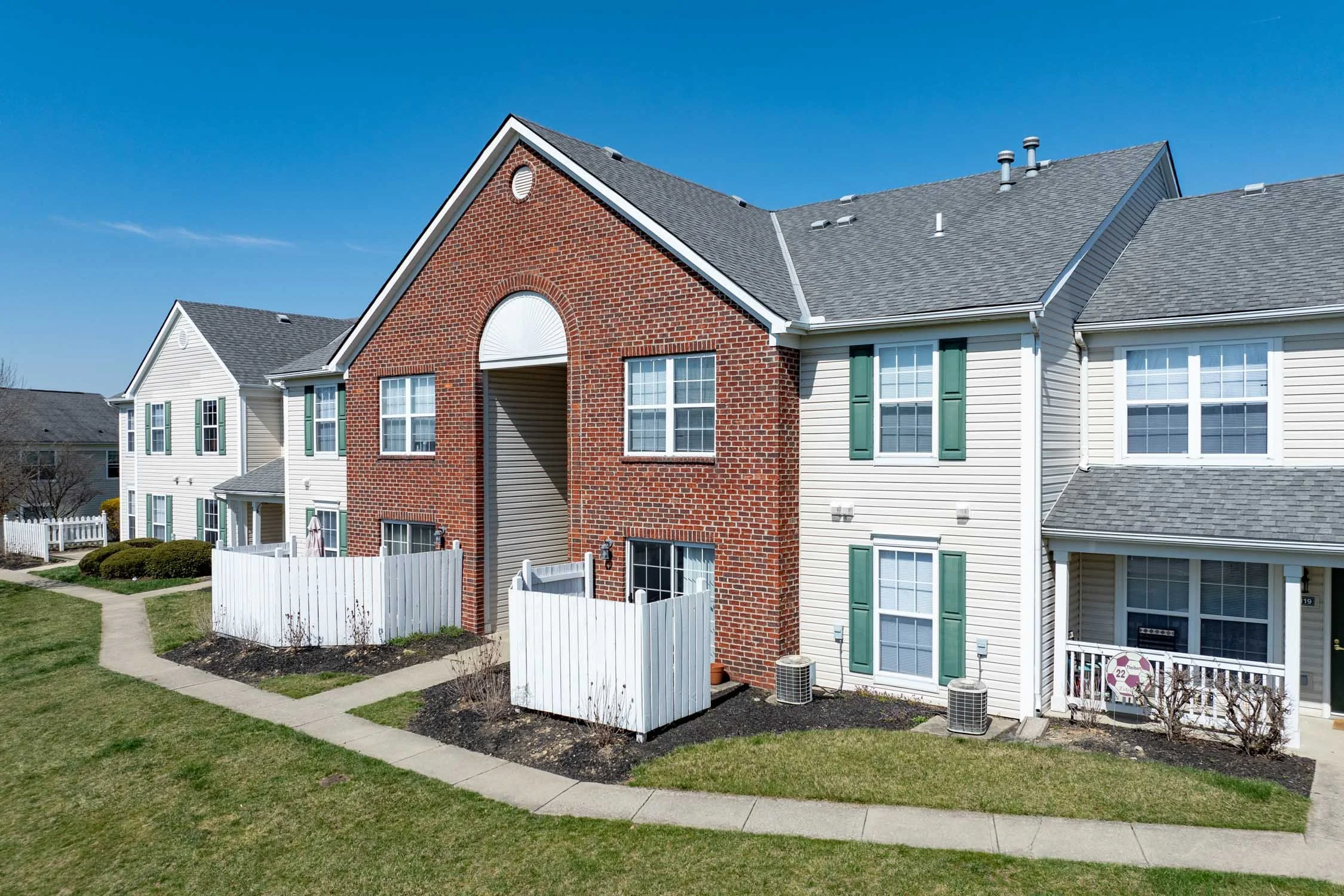 Multi-story residential apartment buildings with brick and siding exteriors, green window shutters, white fences, neatly landscaped lawns, and a clear blue sky.