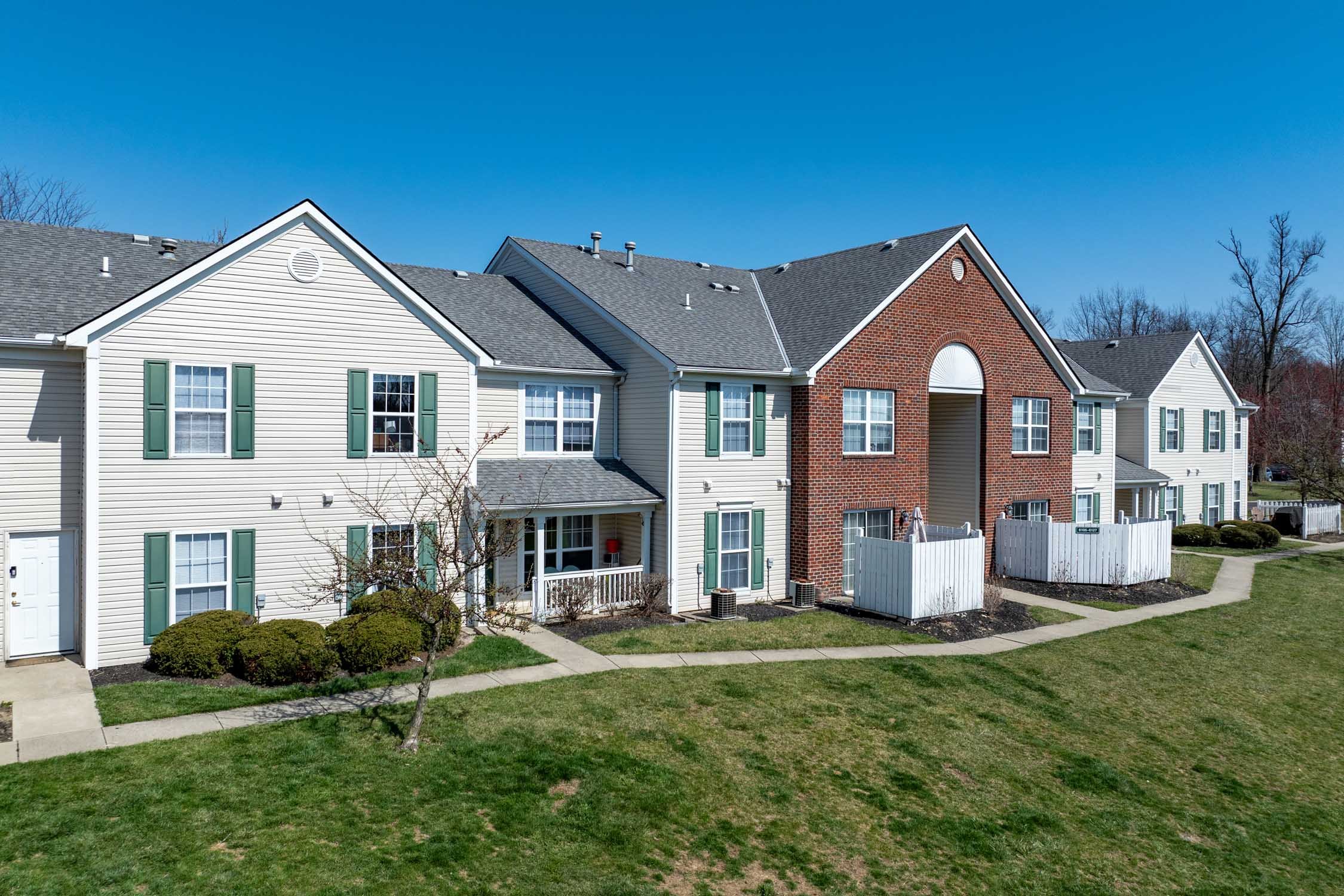 A row of beige and brick townhouses with green shutters, white fences, and small front yards, under a clear blue sky.