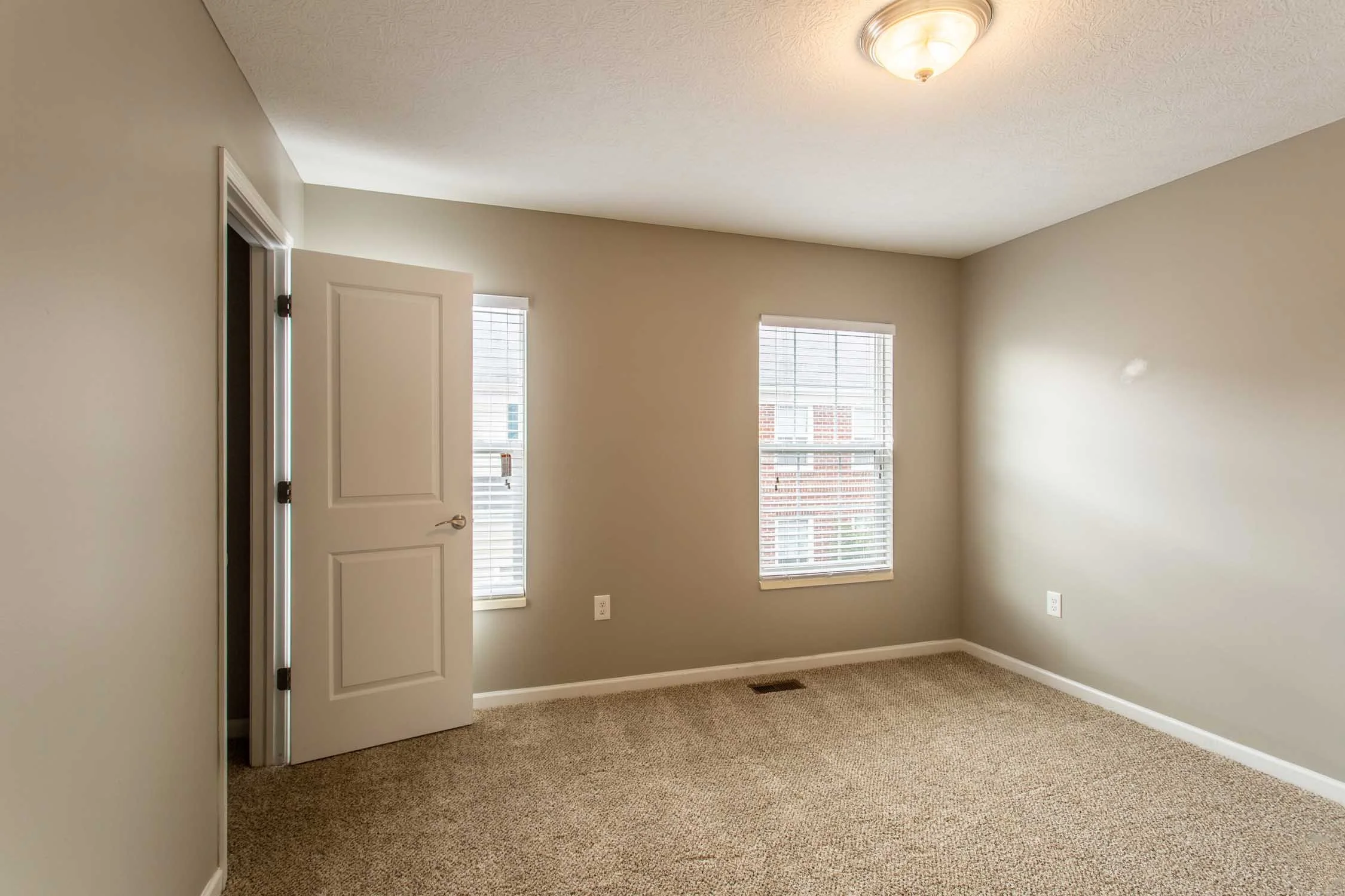 Empty beige-colored room with two windows and a white door partially open, beige carpet, and ceiling light.