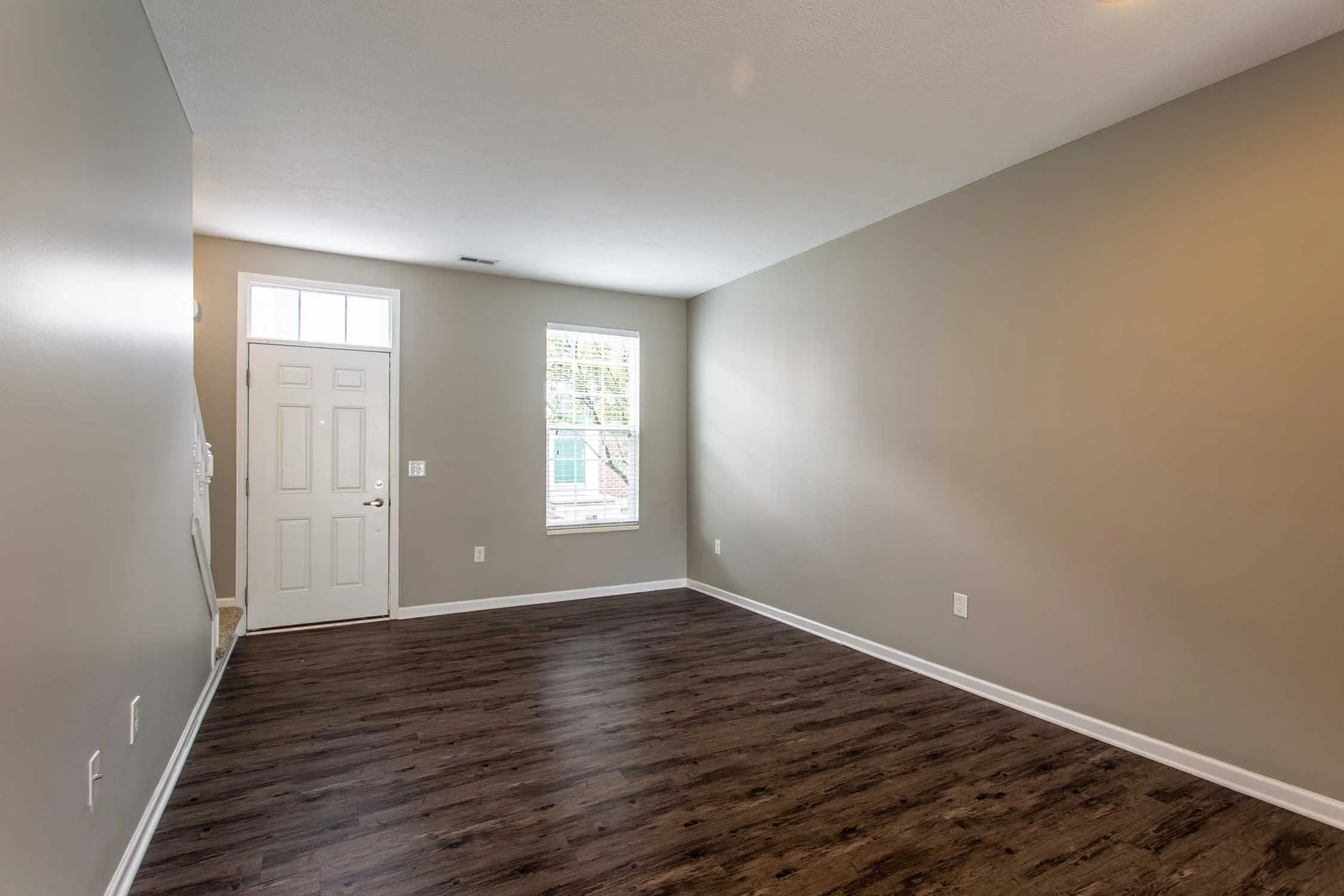 Empty living room with gray walls, white door, windows with blinds, and dark wood flooring.