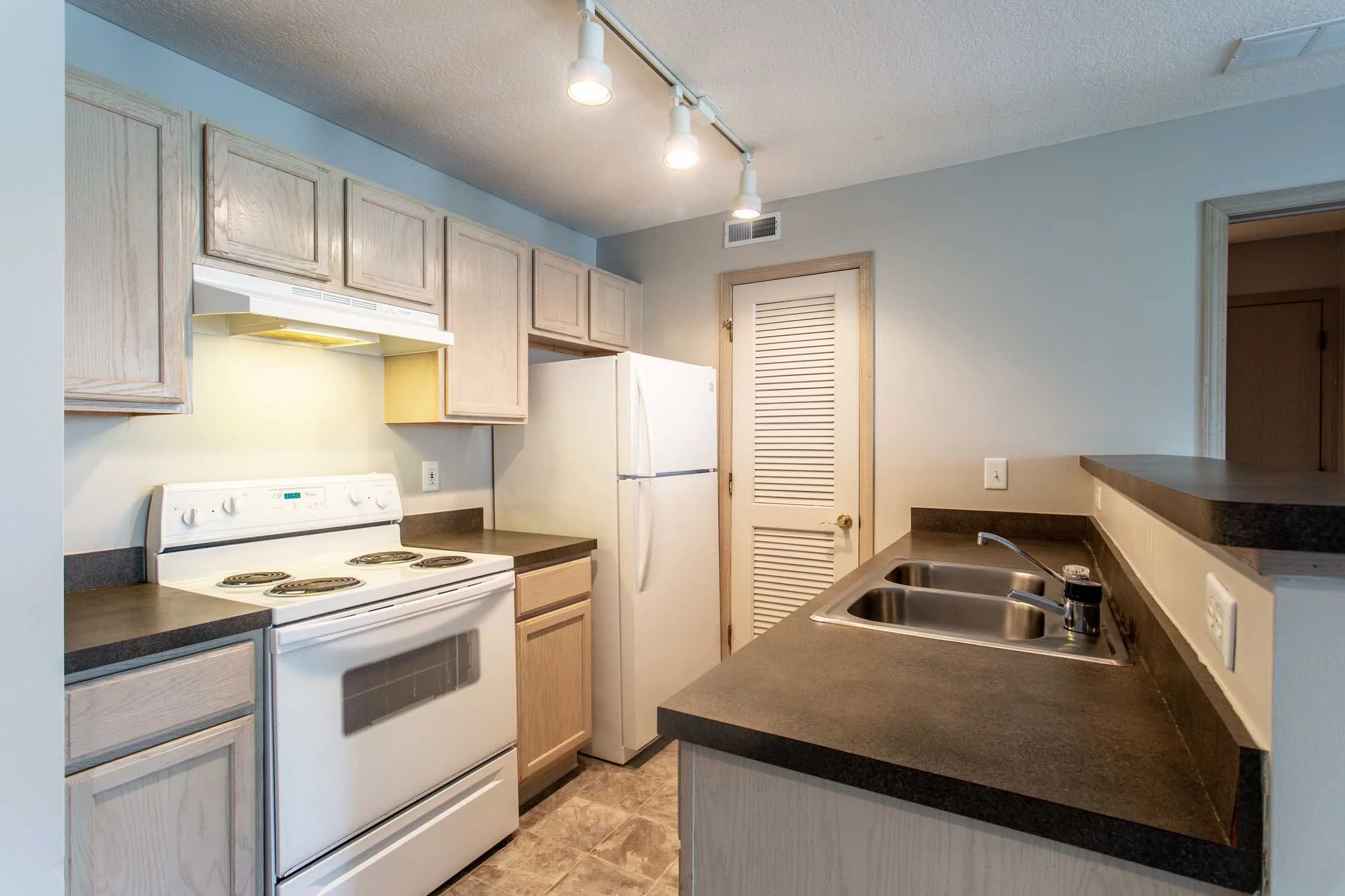 A kitchen with light wood cabinets, white appliances including a stove and refrigerator, a double sink, and dark countertops. Recessed lighting features three lights on a track.