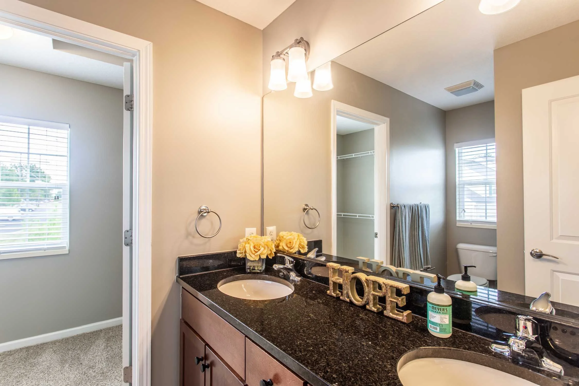 Bathroom interior with a dark granite countertop, sink, decorative flowers, and a mirror. The space has natural light from windows, and there are wall-mounted towel rings and a white cabinet. The decor includes a sign spelling 'hope' and a bottle of 