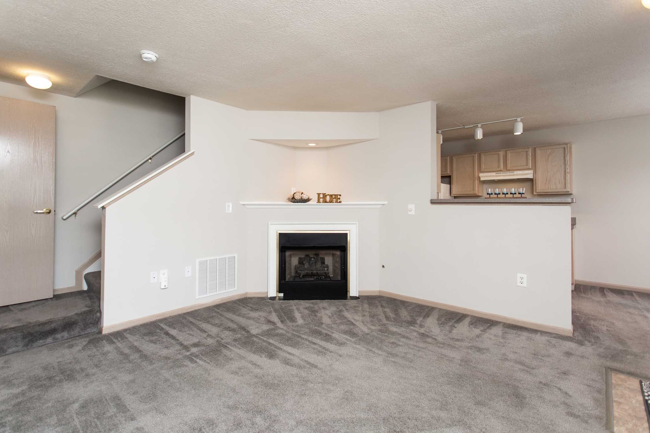 Living room with a gas fireplace, mantel, and decorative items that say 'HOPE', with stairs leading up on the left and kitchen cabinets to the right, carpeted flooring, and ceiling lights.