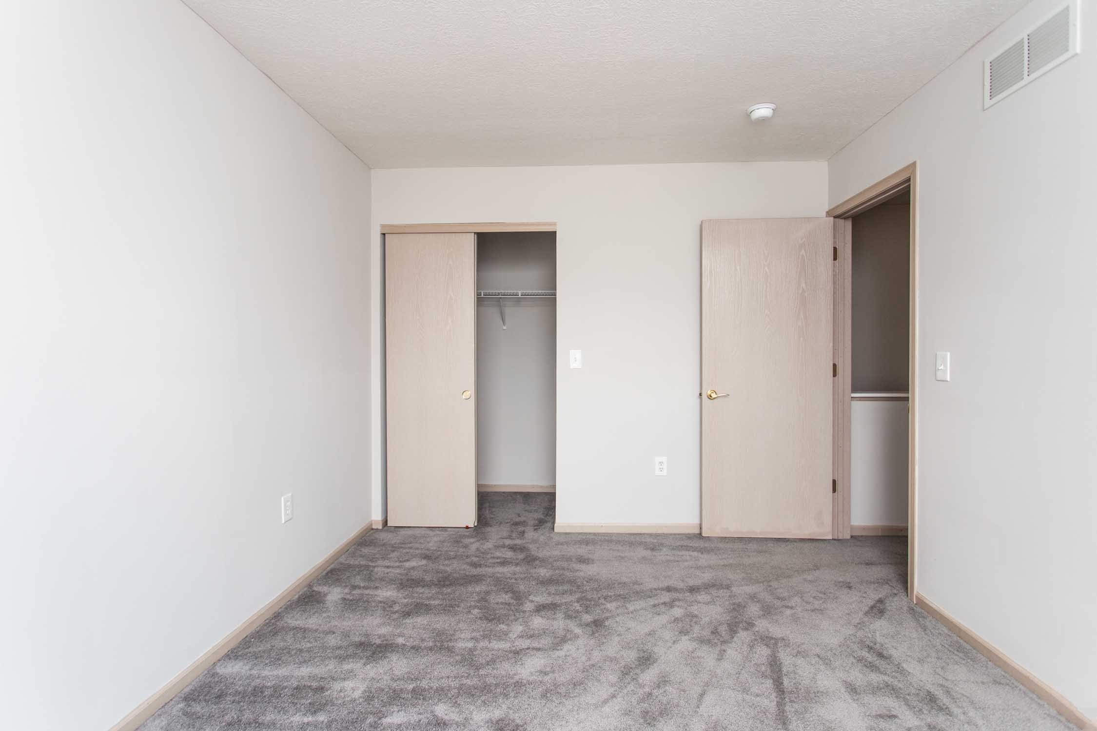 Empty room with gray carpet, white walls, and two open closet doors with a small closet space inside. There is a closed door next to the closet, and ceiling with a smoke detector and air vent.