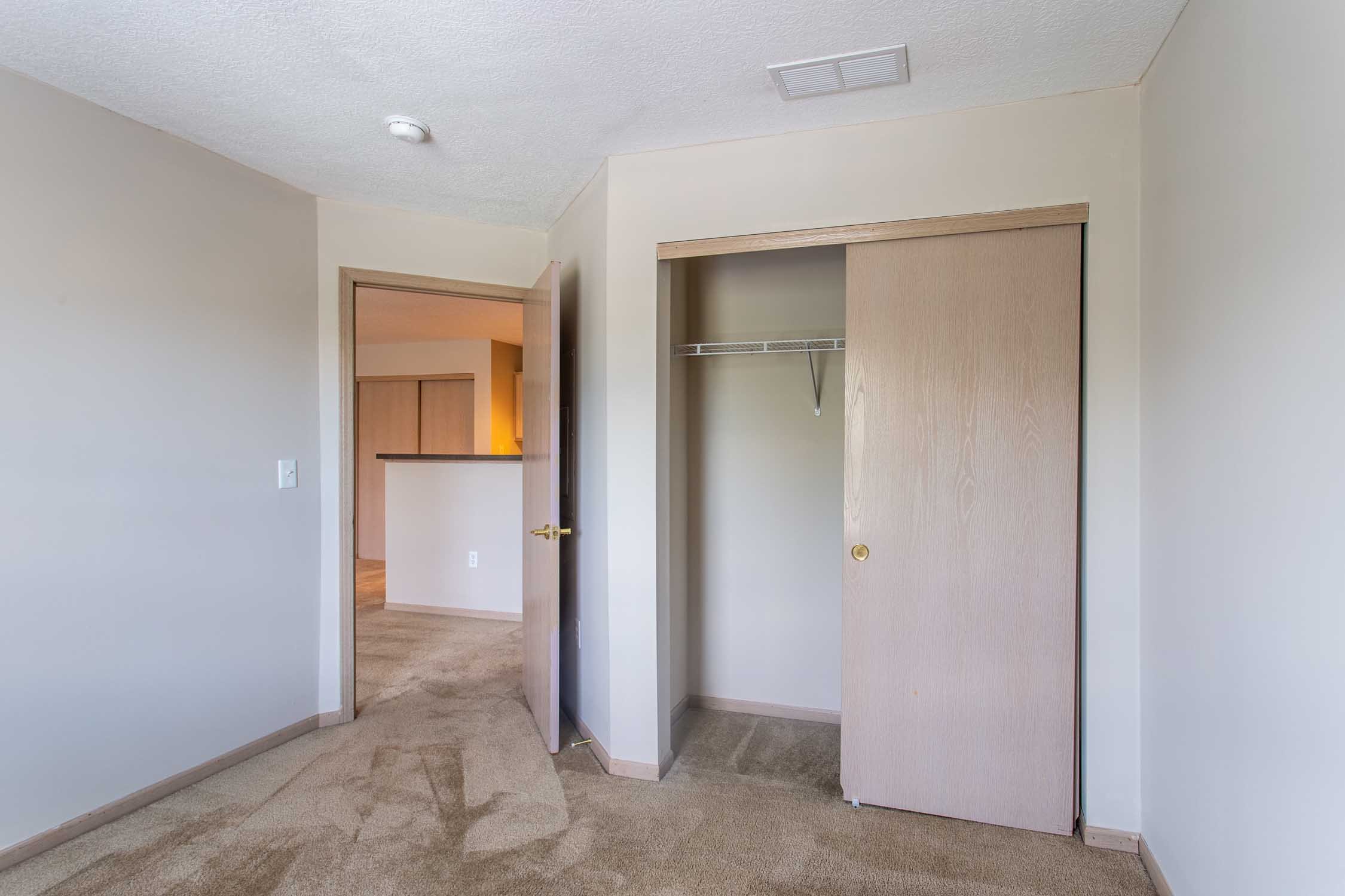 Interior view of a room with beige carpet, open closet with sliding door, and a doorway leading to another room, ceiling vent, and smoke detector.
