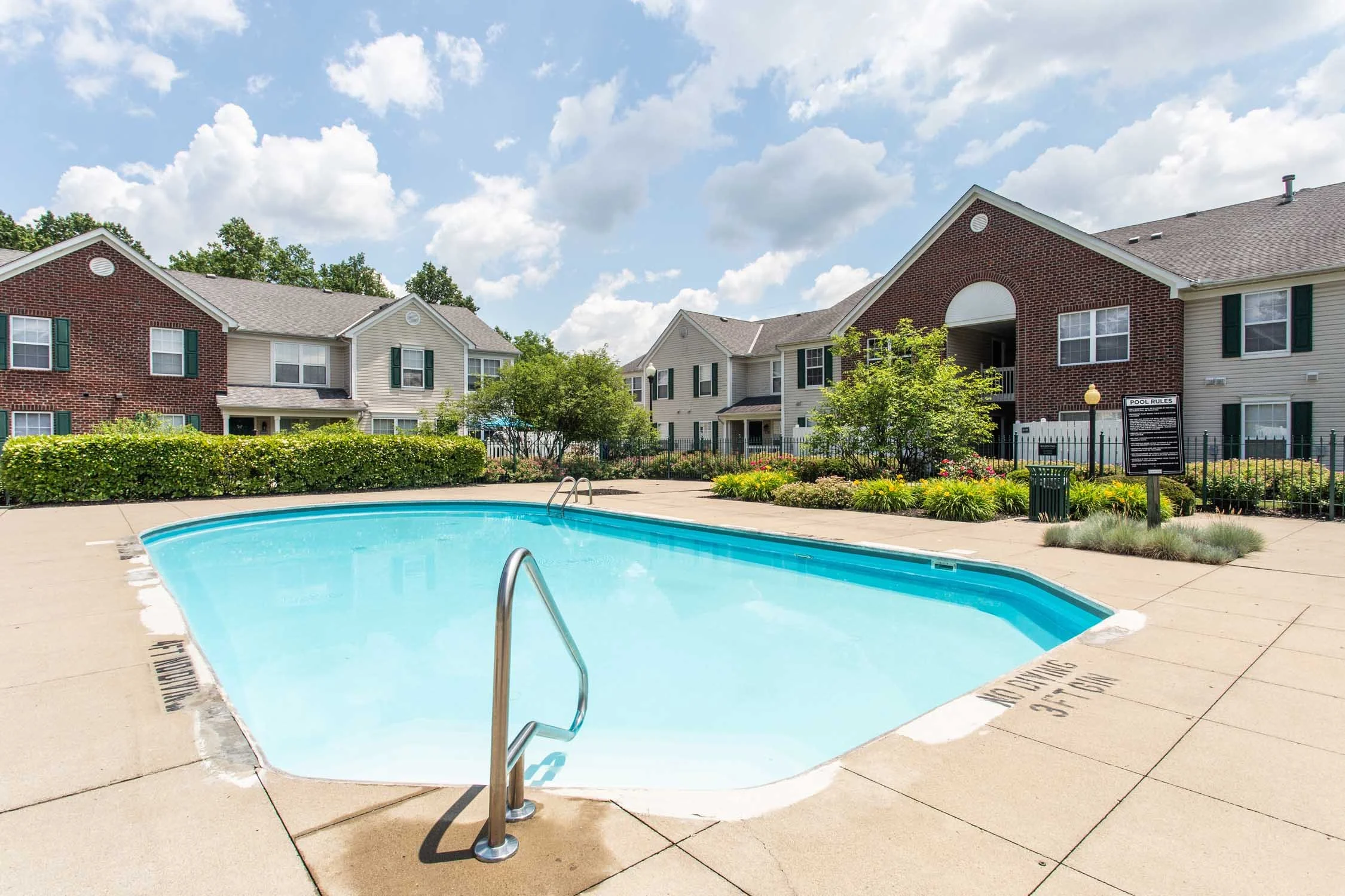 An outdoor swimming pool surrounded by a concrete deck, with residential apartment buildings in the background, lush green trees, and a partly cloudy blue sky.
