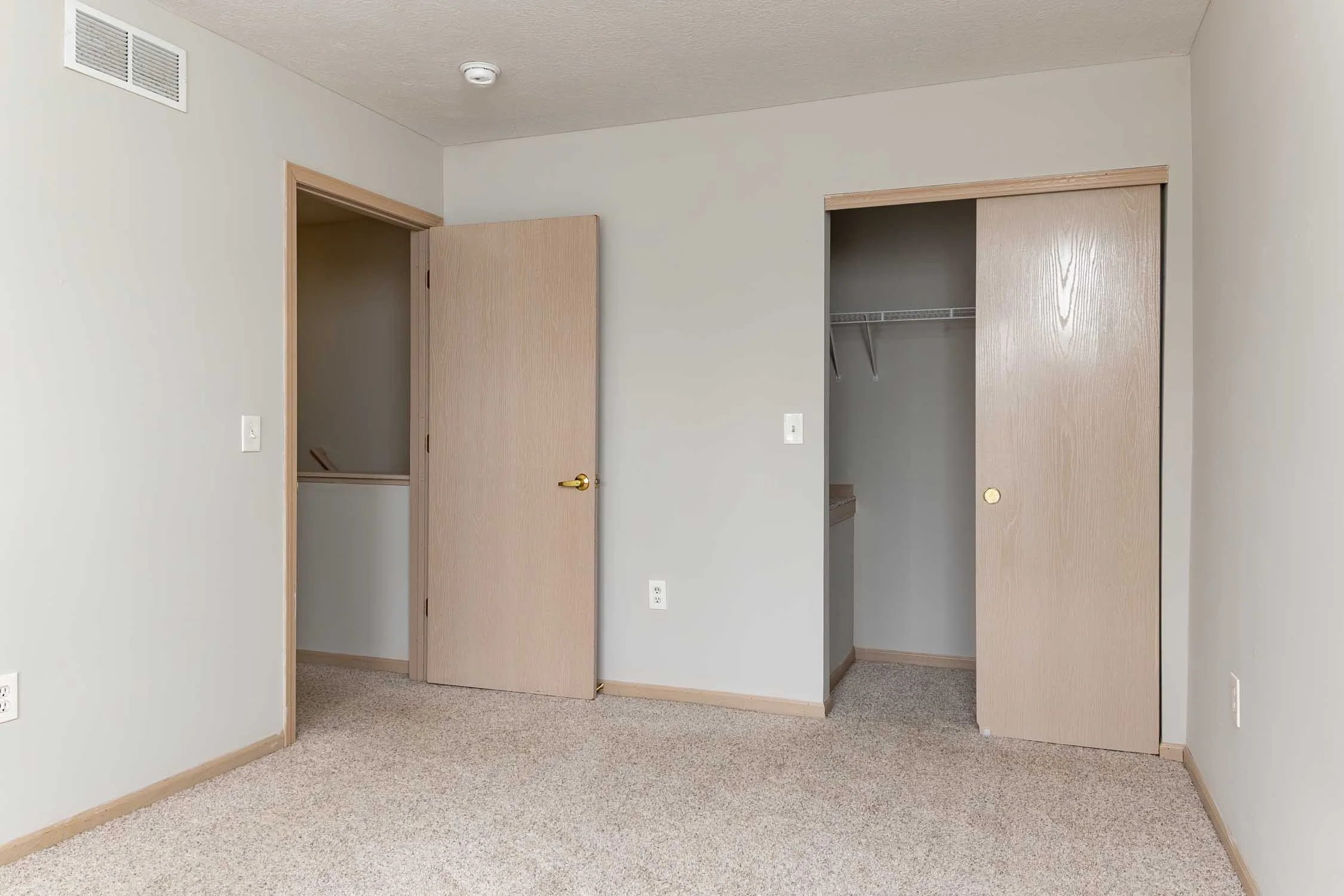 Empty room with beige carpet and light-colored walls, featuring two open closet doors and a plain door, with a smoke detector on the ceiling.