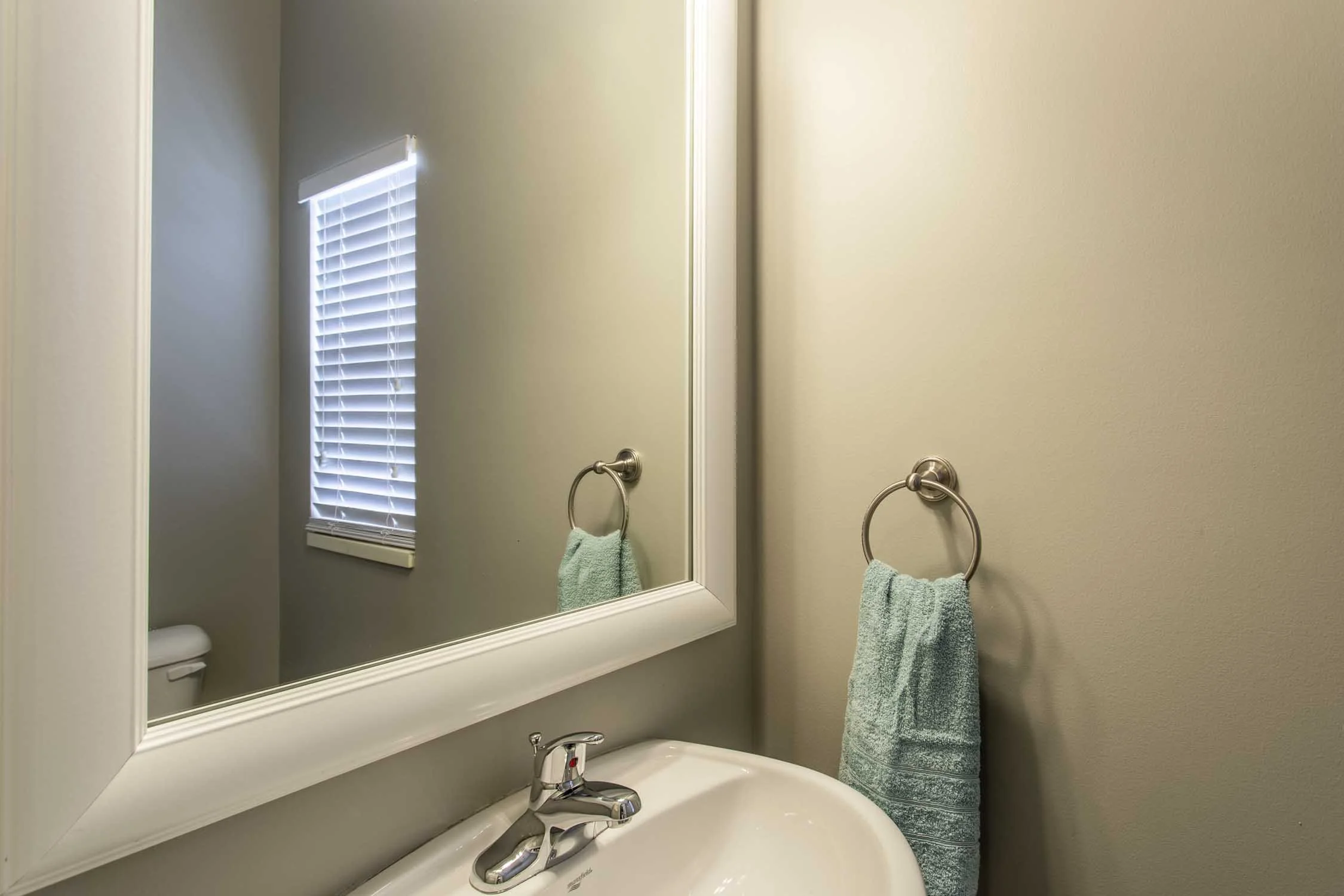 A bathroom sink with a mirror above it, a towel ring with a green towel, and a window with blinds reflected in the mirror.
