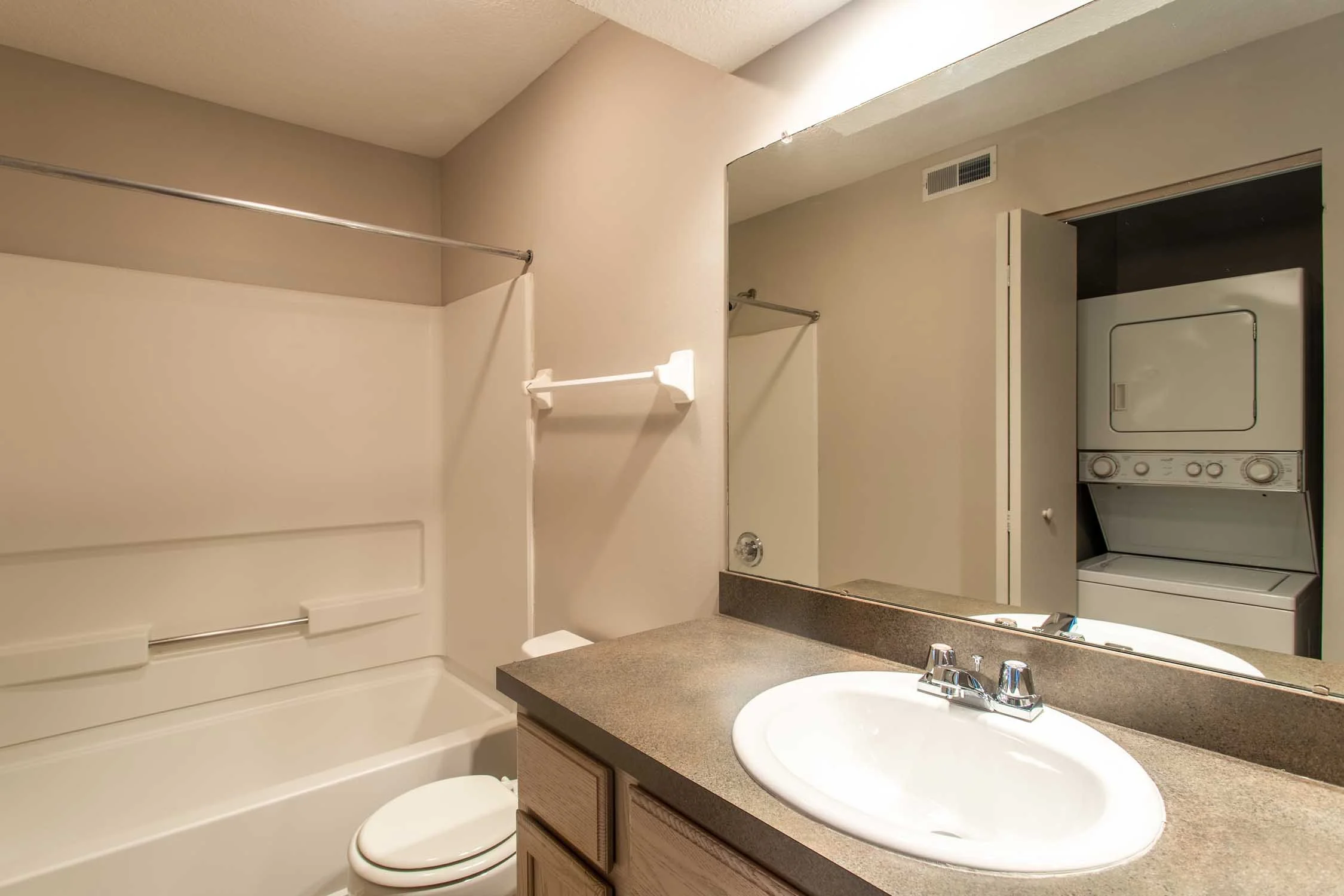 A bathroom with a bathtub, toilet, and vanity sink. There is a mirror above the sink, beige countertops, and a washer and dryer stacked in a closet space.