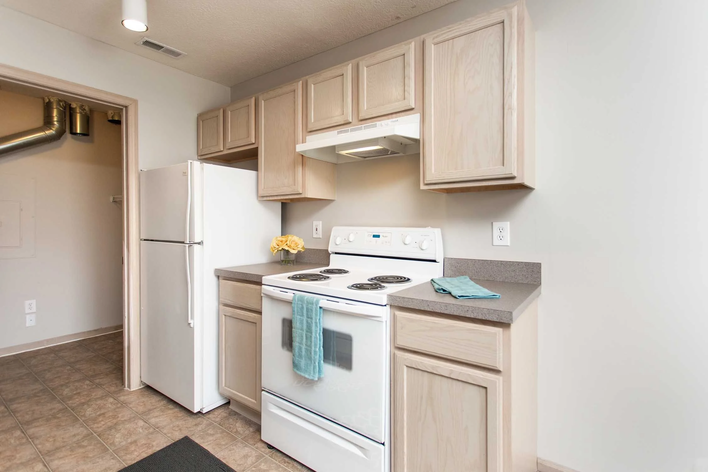 Kitchen with light wood cabinets, white refrigerator, white stove with oven, gray countertop, small vase with yellow flowers, and two folded blue towels hanging on the oven handle. The floor is tiled, and there are two electrical outlets on the wall.