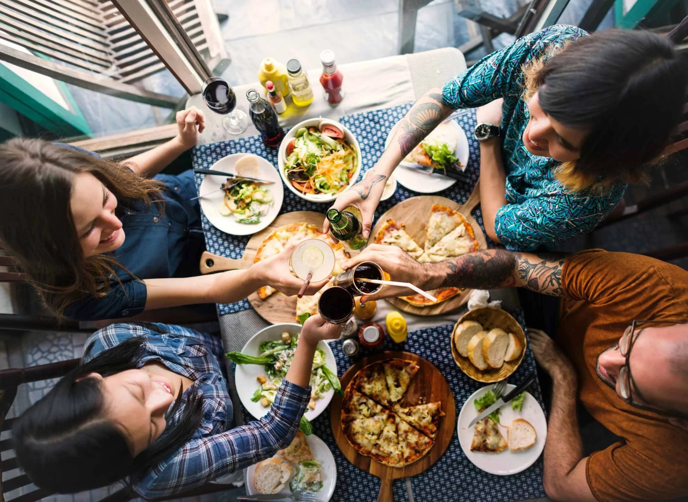 People sharing a meal at a table with salad, pizza, bread, salads, and drinks, outdoors near a glass door.