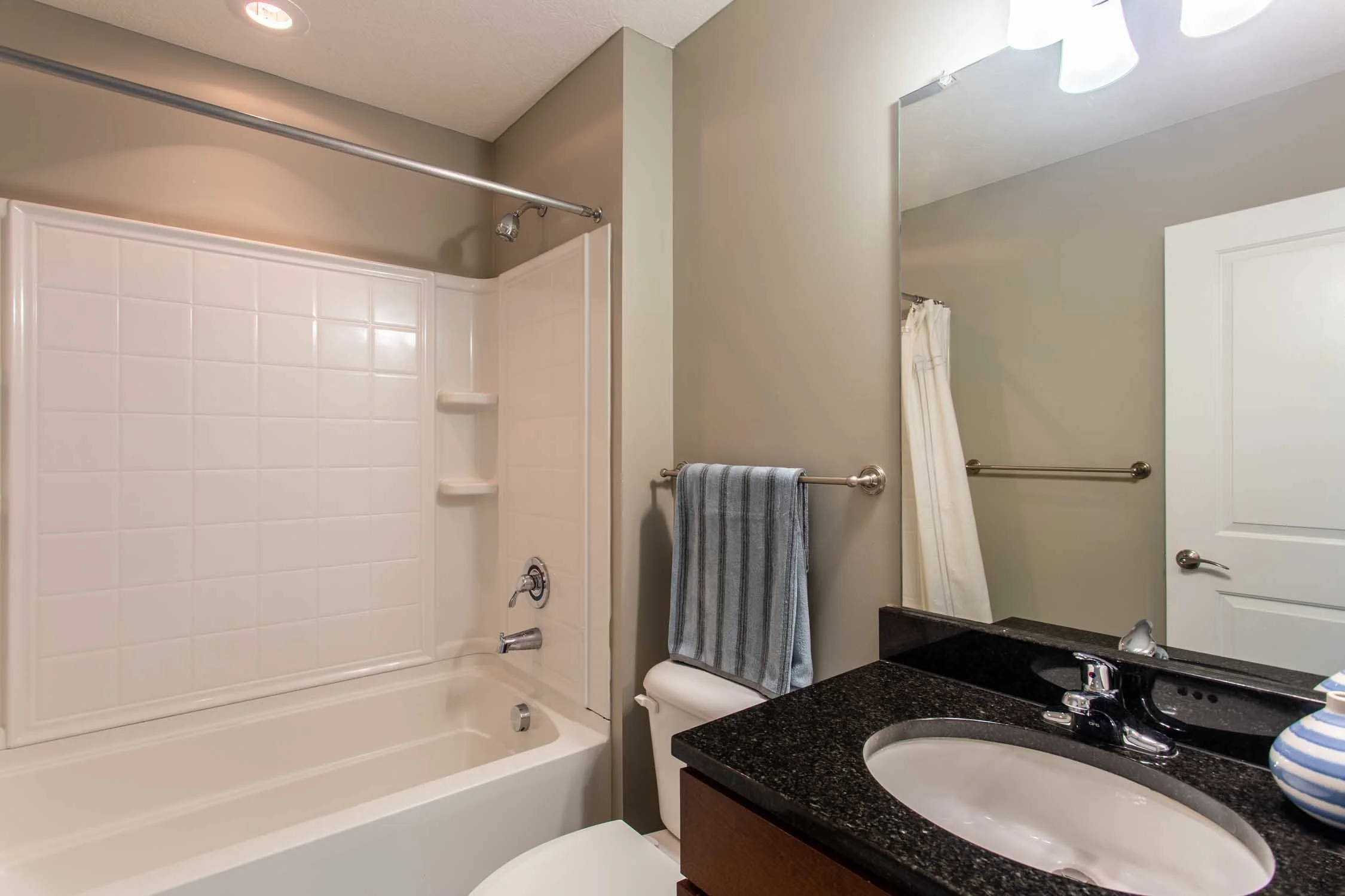 Bathroom shower with white tile surround, gray walls, a towel hanging on a towel bar, a mirror, and a black granite sink countertop.