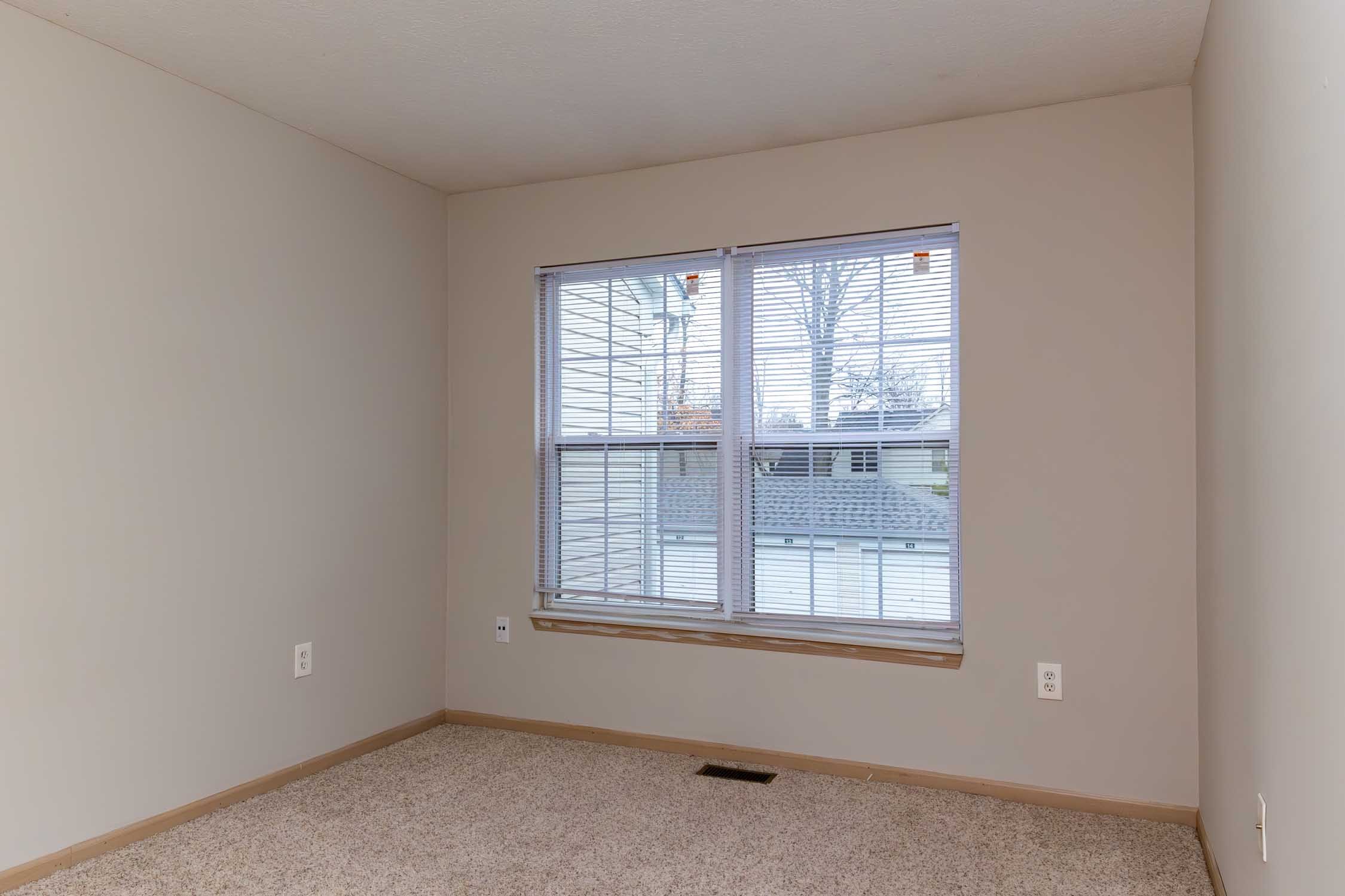 Empty room with beige walls, beige carpet, and a window with blinds showing a view of leafless trees outside.