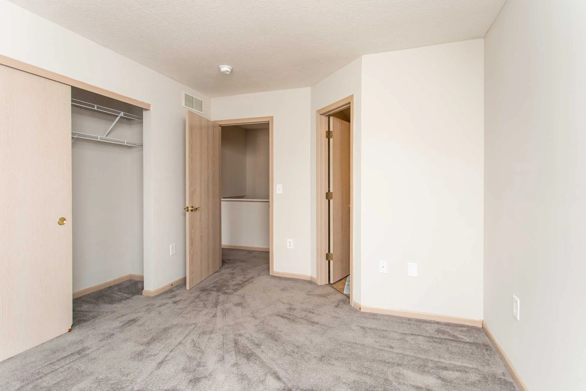Empty bedroom with light-colored walls, brown carpet, open closet with wire shelves, and doorways leading to other rooms.