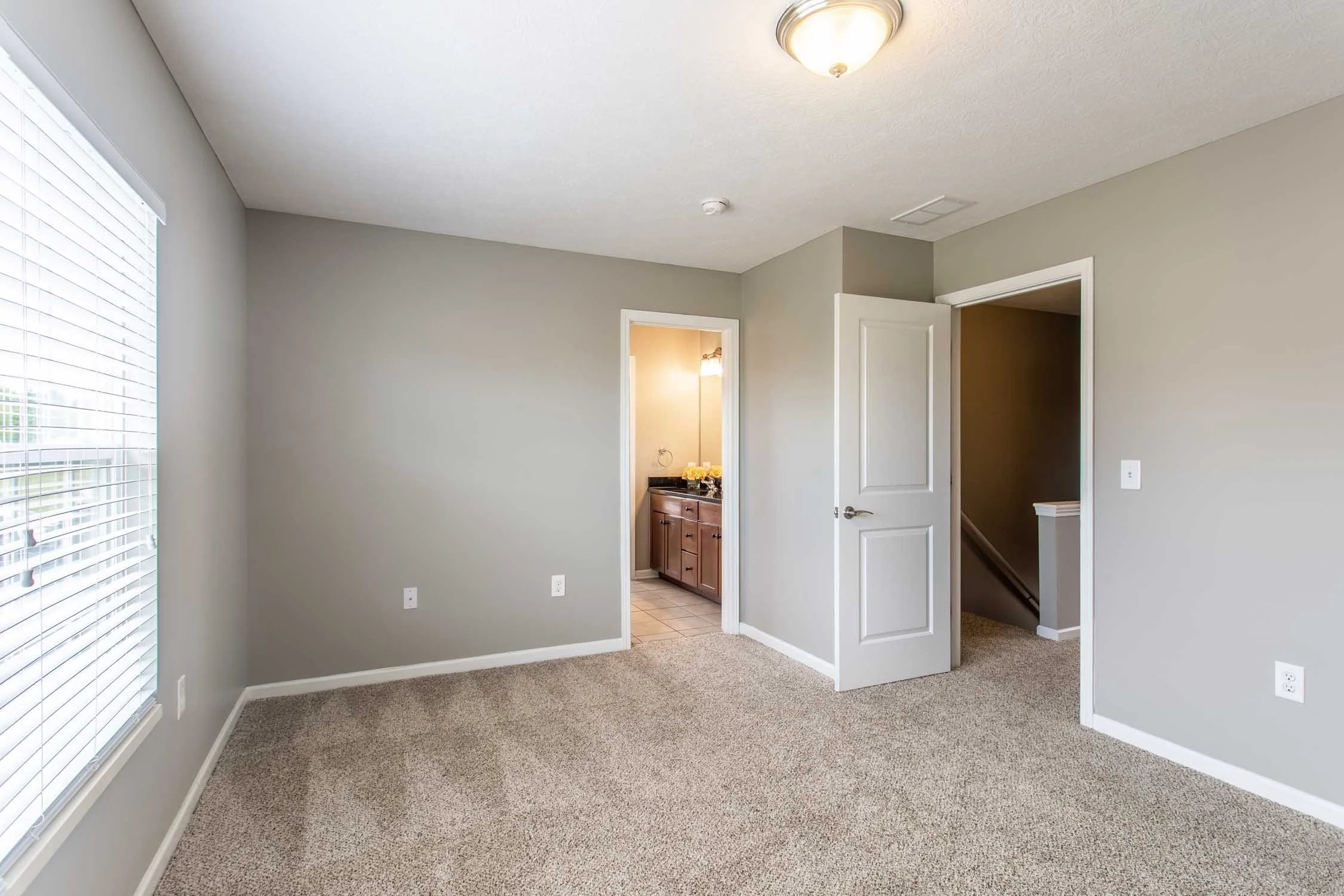 A room with beige carpet, light gray walls, a large window with white blinds, an open door leading to a bathroom with a wooden vanity and a flower arrangement, and another door leading to a stairway.