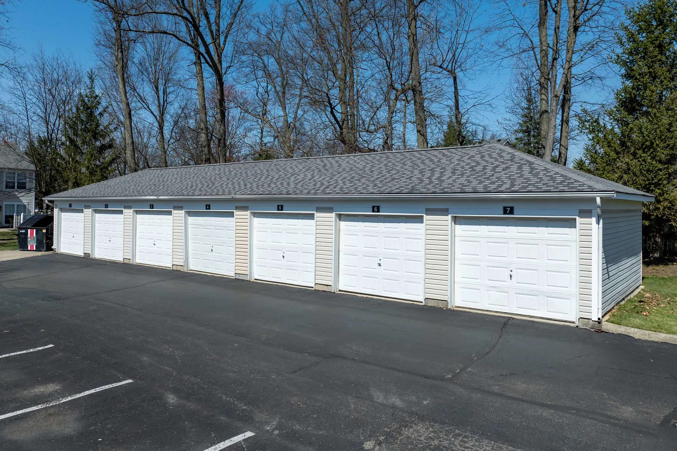 A row of six white garage doors numbered 1 through 7, part of a small building, with a parking lot in front and leafless trees in the background on a clear day.