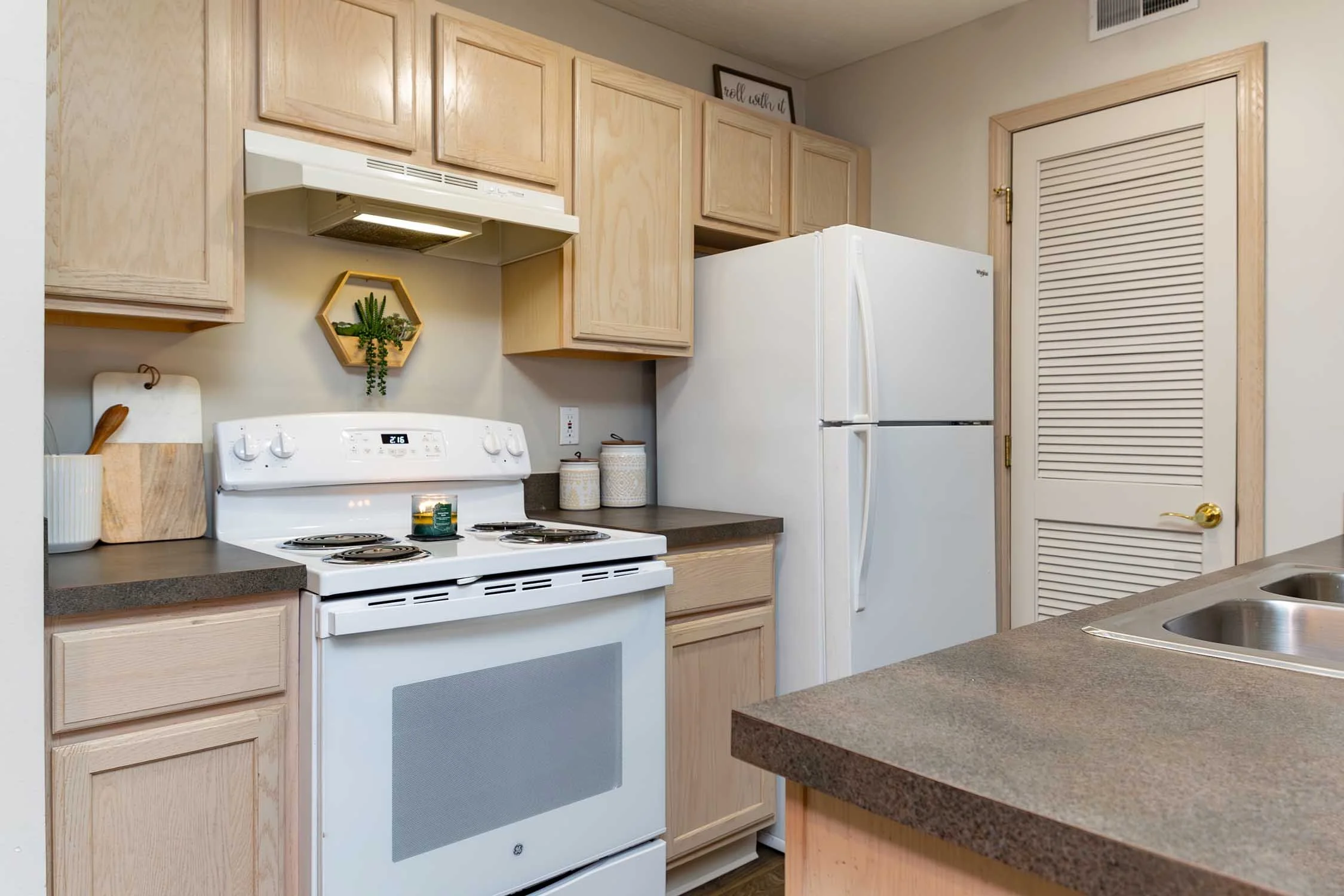 A kitchen with light wood cabinets, a white stove, a white refrigerator, a beige countertop, and a double sink. There are decorative items including a small shelf with plants, a cutting board, and containers. A framed sign is on top of the cabinets, 