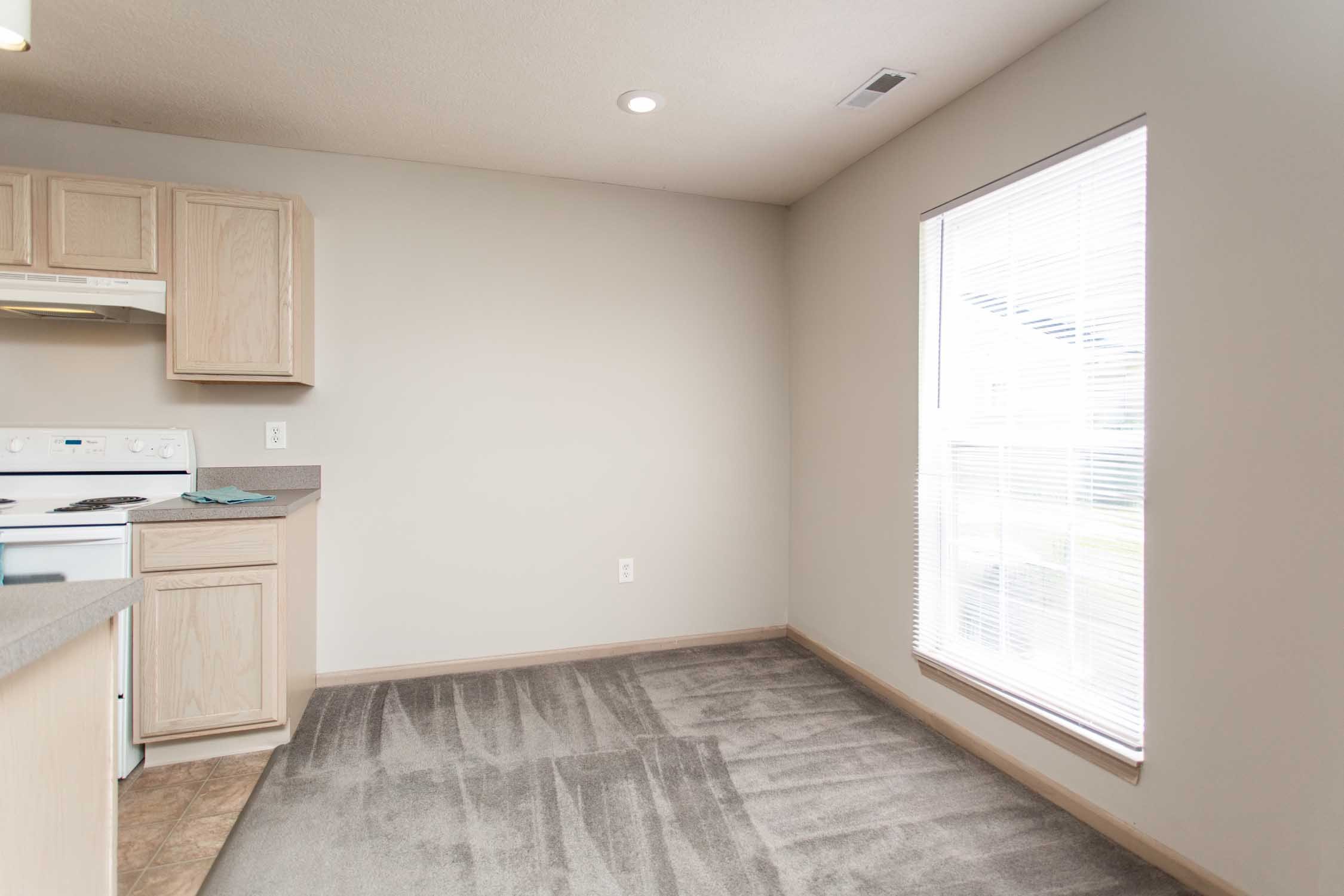 A corner of a room with a large window with blinds, beige walls, wooden cabinetry, a white stove, and a gray carpeted floor.