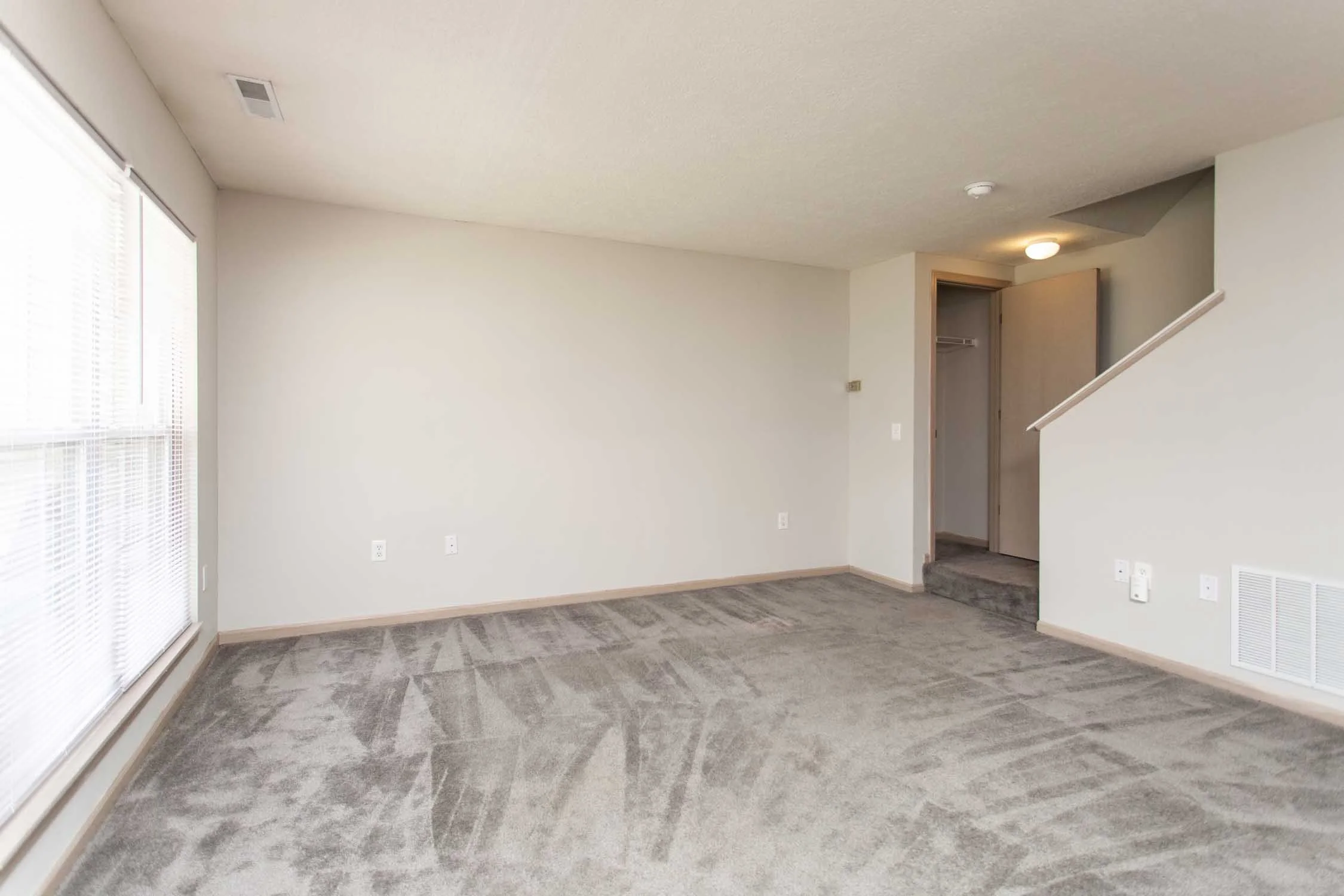 Empty living room with beige walls, large window with blinds, gray carpet, and an open doorway leading to a closet and stairs.