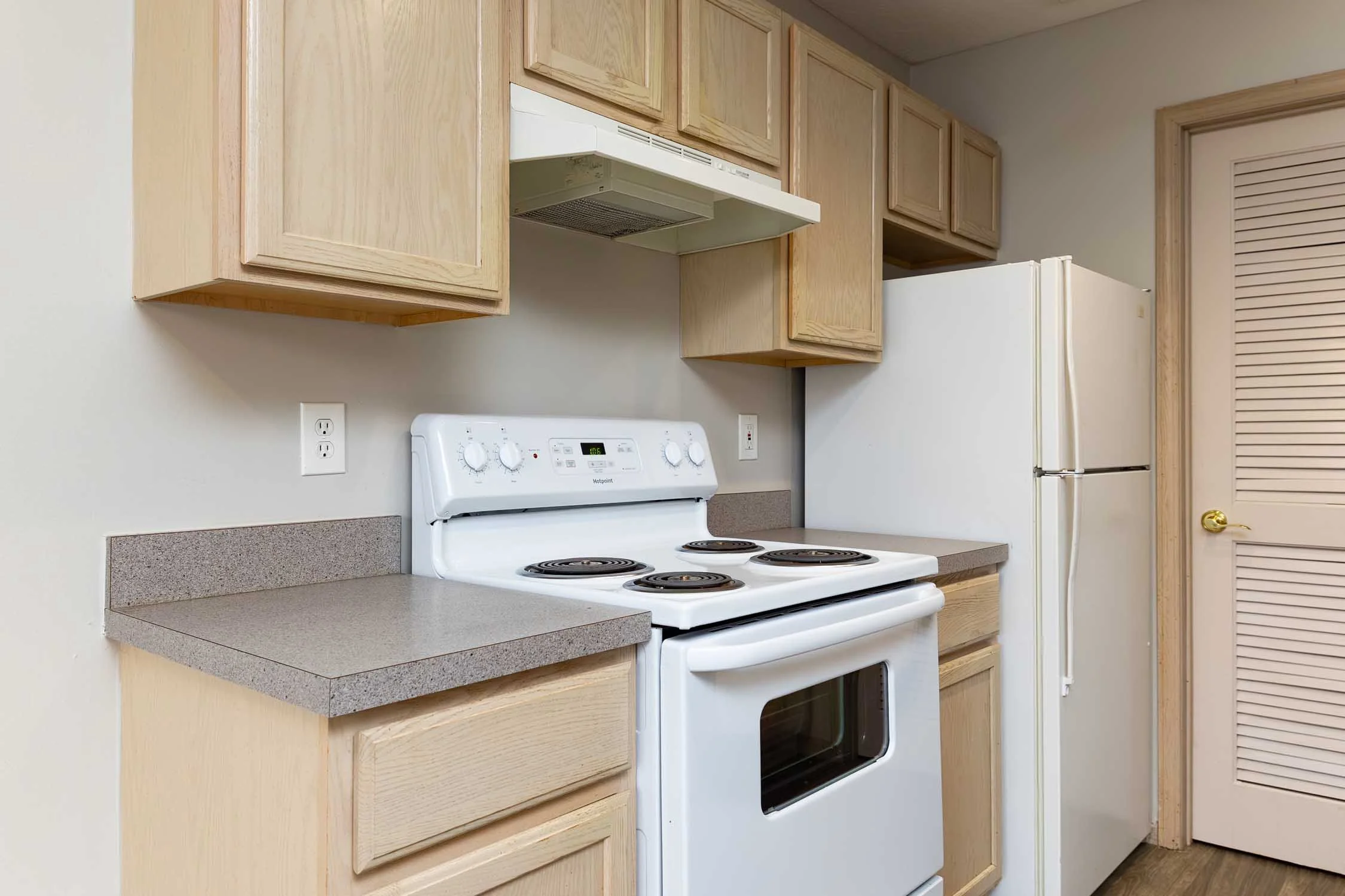A kitchen with light wood cabinets, a white stove with coil burners, a white refrigerator, and a doorway with a louvered door.