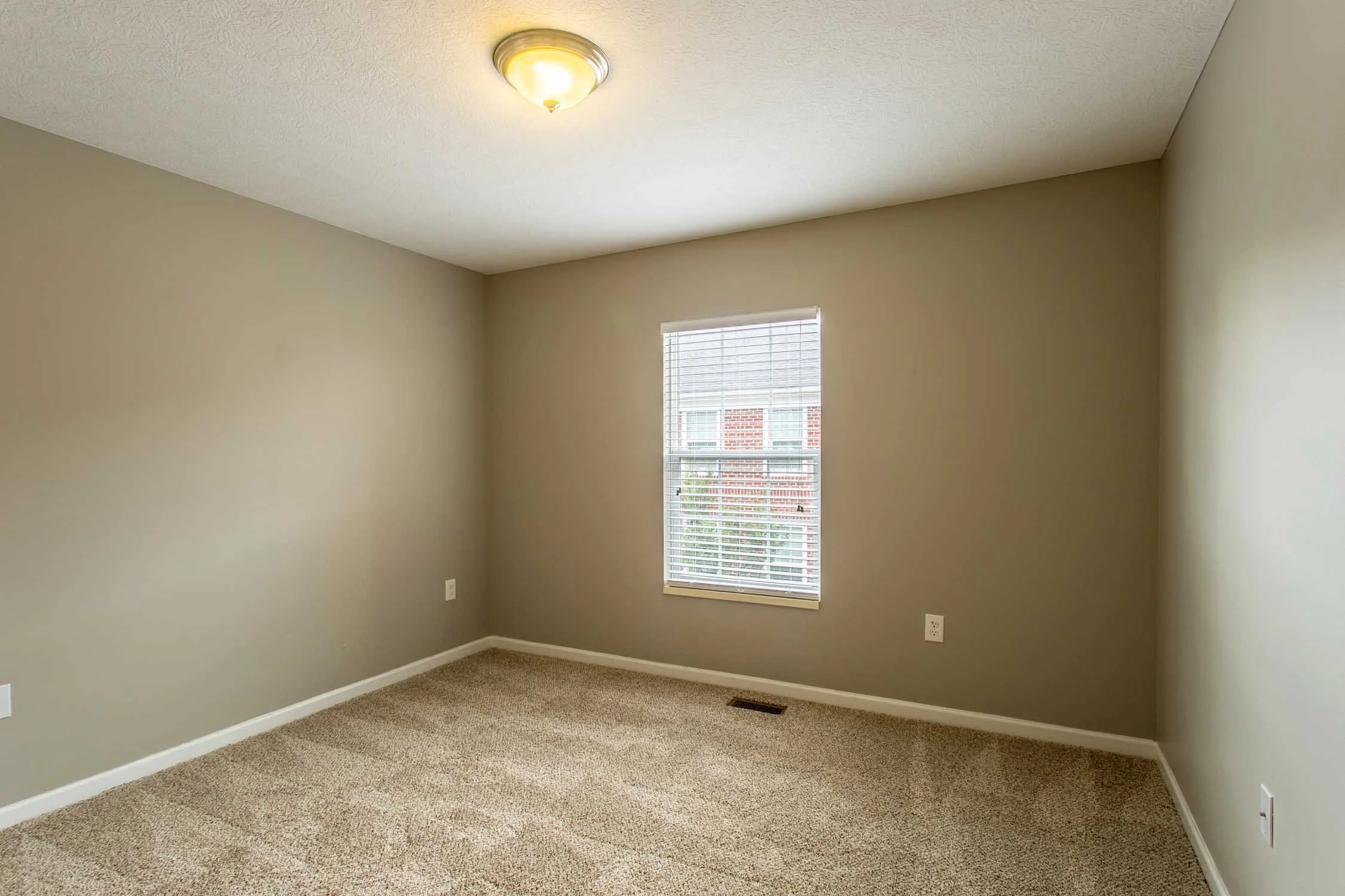 Empty room with beige walls, a single window with blinds, carpeted floor, ceiling light fixture, and electrical outlets.