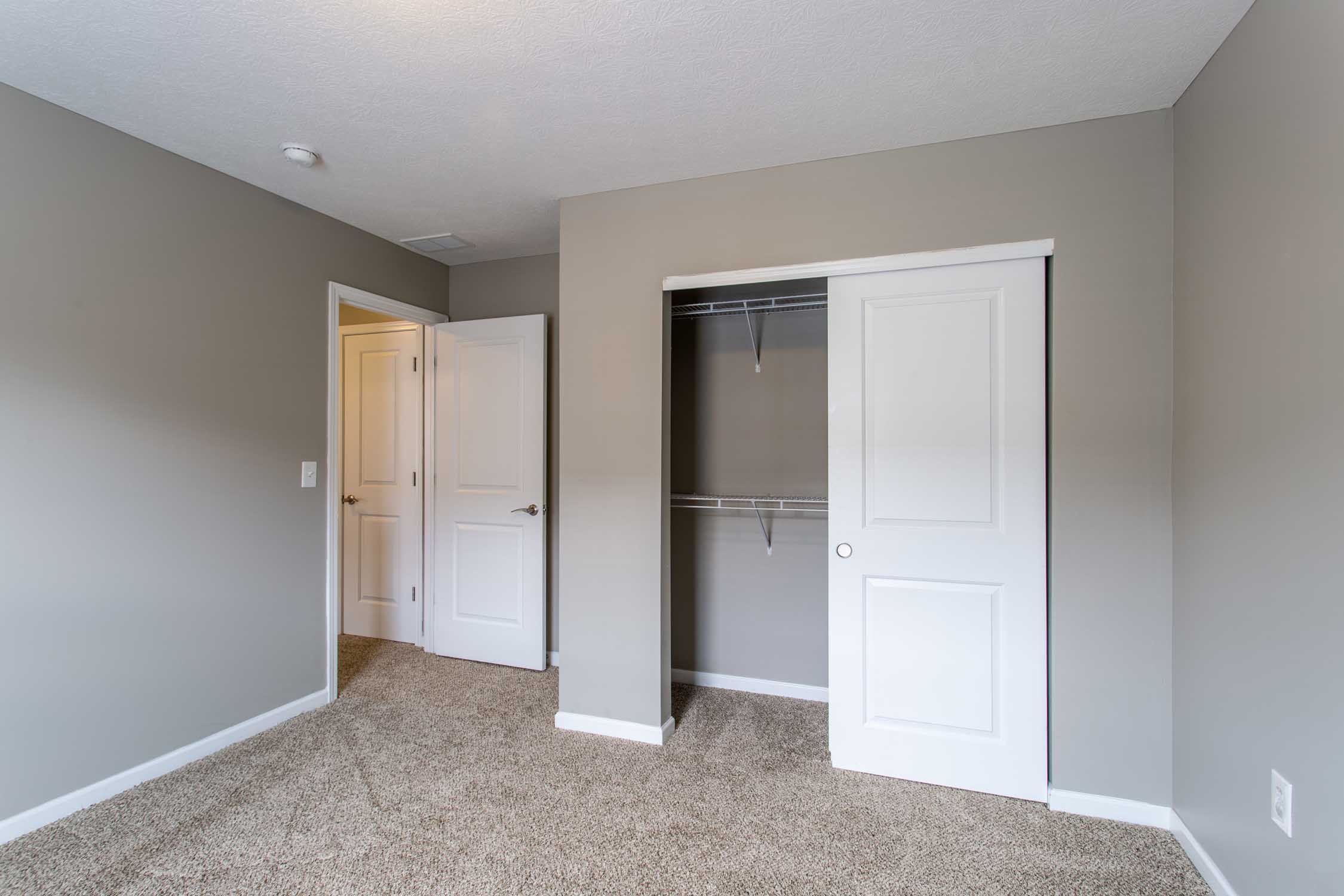 Empty bedroom with beige carpet, gray walls, open closet with white sliding doors, and a white door partially open leading to another room.