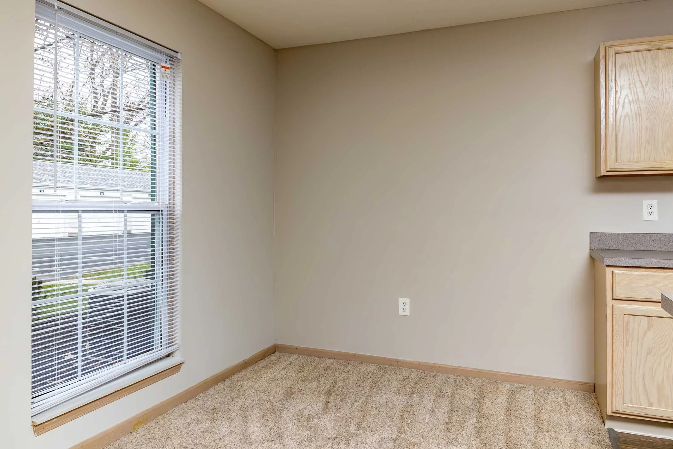 Empty kitchen corner with a window with blinds, beige walls, beige carpet, and light wood cabinetry.