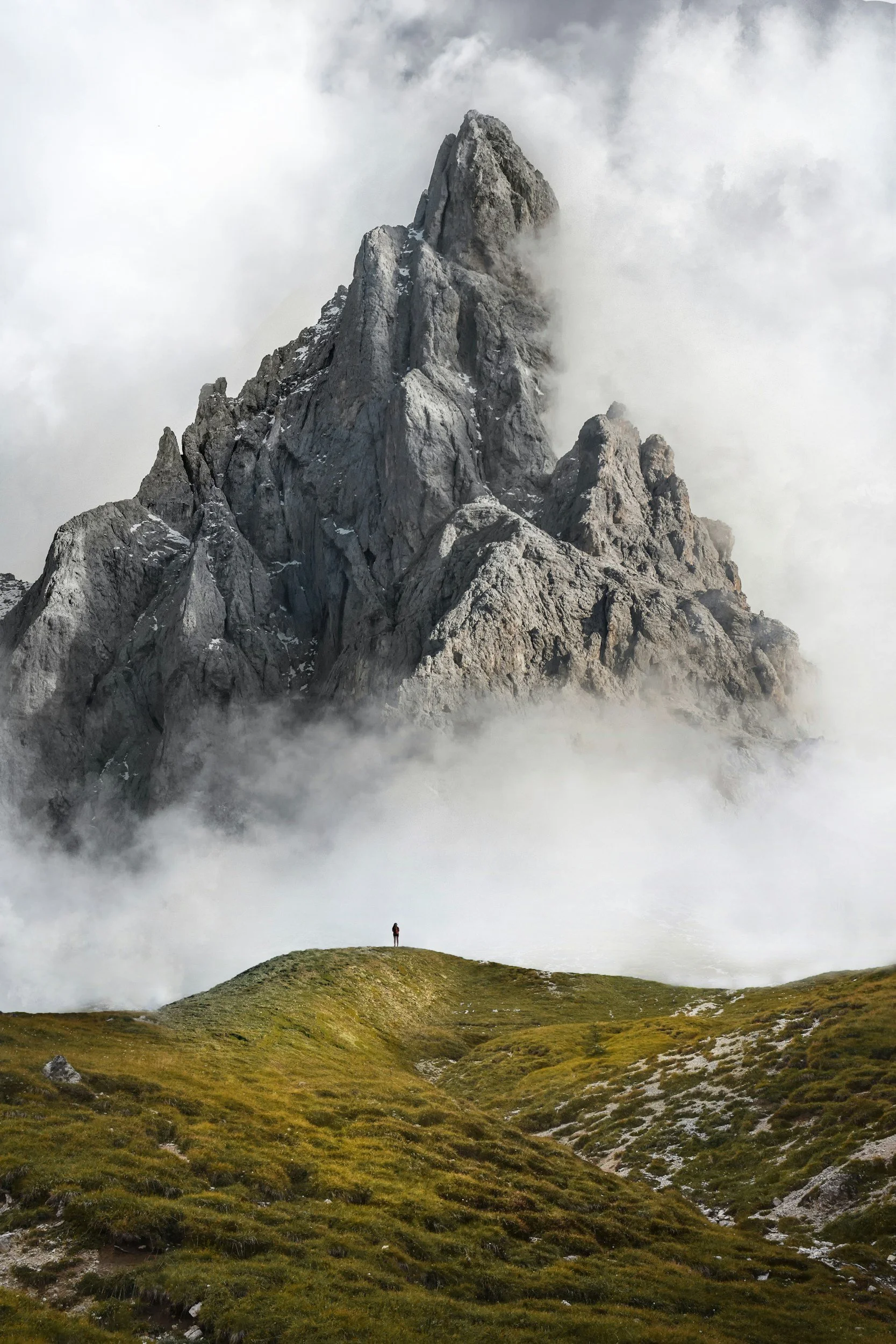 A lone person standing on a grassy hilltop in front of a dramatic mountain peak partially shrouded in clouds and mist.