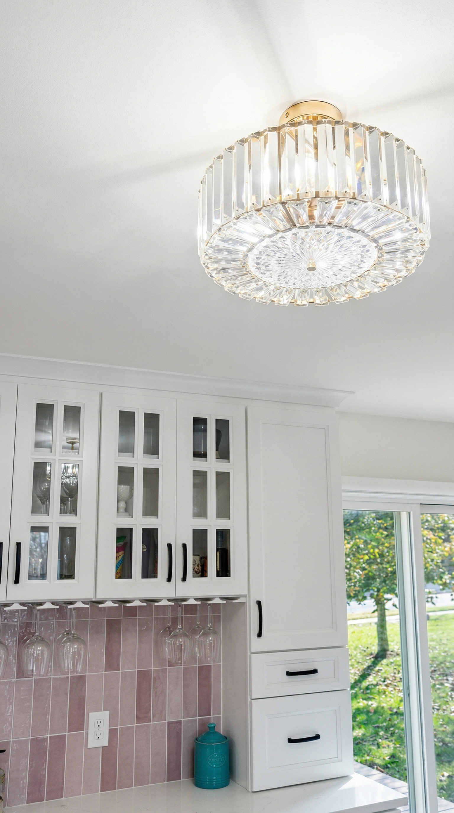 A white kitchen with a chandelier and glass cabinet doors, a window showing trees outside, pink tiles on the backsplash, and a blue jar on the floor.