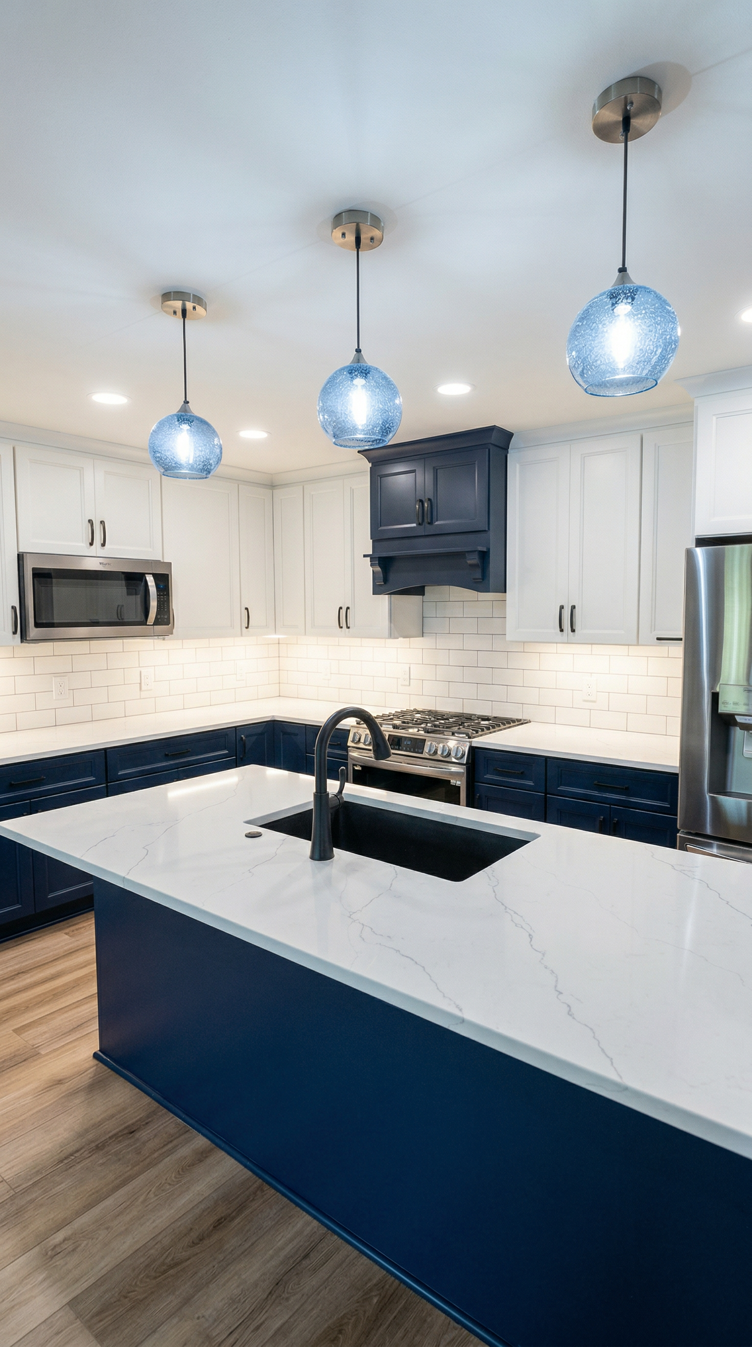 Modern kitchen with a white island featuring a black sink, white and navy blue cabinetry, stainless steel appliances, white subway tile backsplash, and three blue pendant lights hanging from the ceiling.