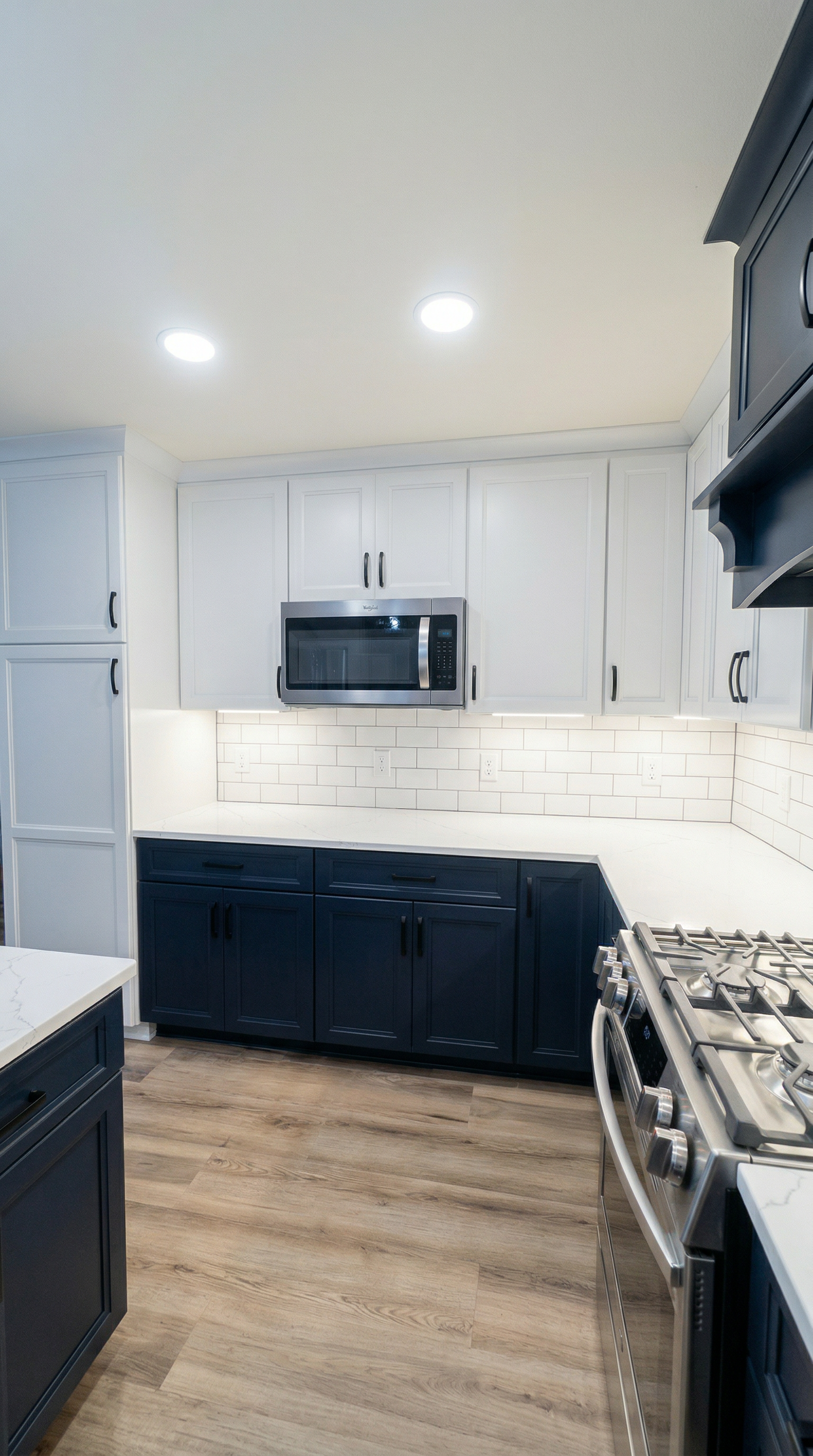Modern kitchen with white upper cabinets, navy lower cabinets, white countertops, a stainless steel microwave oven above the counter, a gas stove on the right, and a white subway tile backsplash. Recessed ceiling lights illuminate the space, and ther