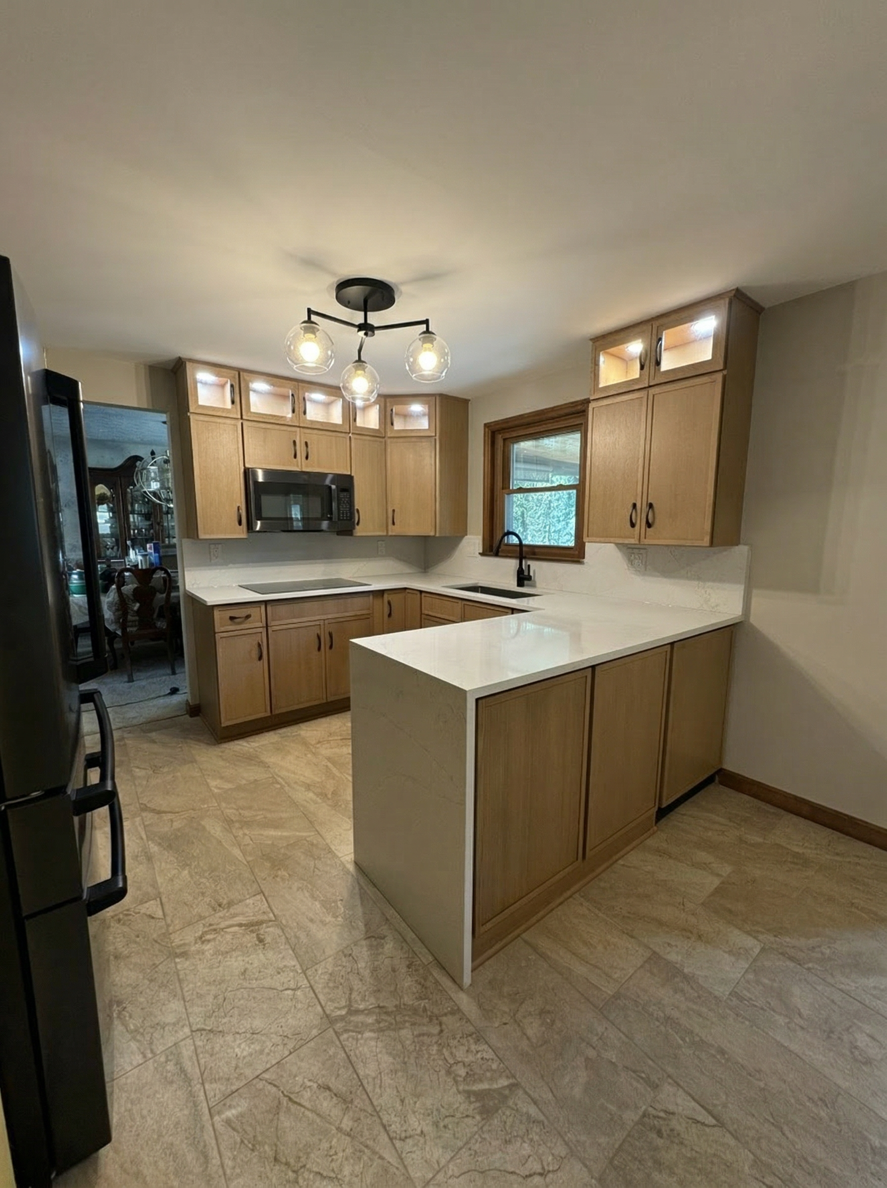 Kitchen with light wood cabinets, black sink, microwave, and partially visible dining area with a table and chairs. Tile flooring and ceiling light fixture with four bulbs and glass globes.