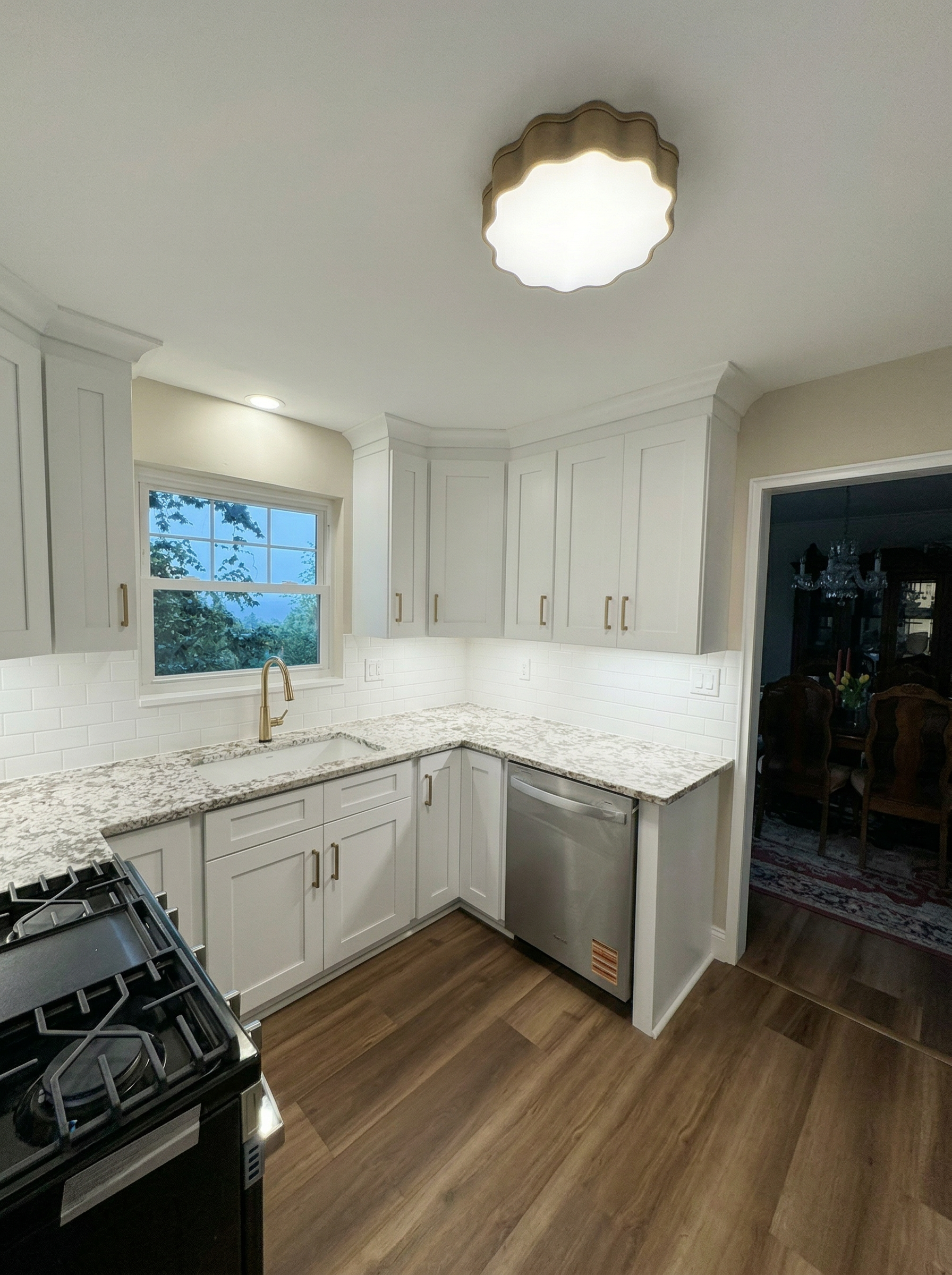 Modern kitchen with white cabinets, granite countertops, a window with trees outside, and built-in appliances, hardwood floors, and a doorway leading to a dining area with a chandelier.