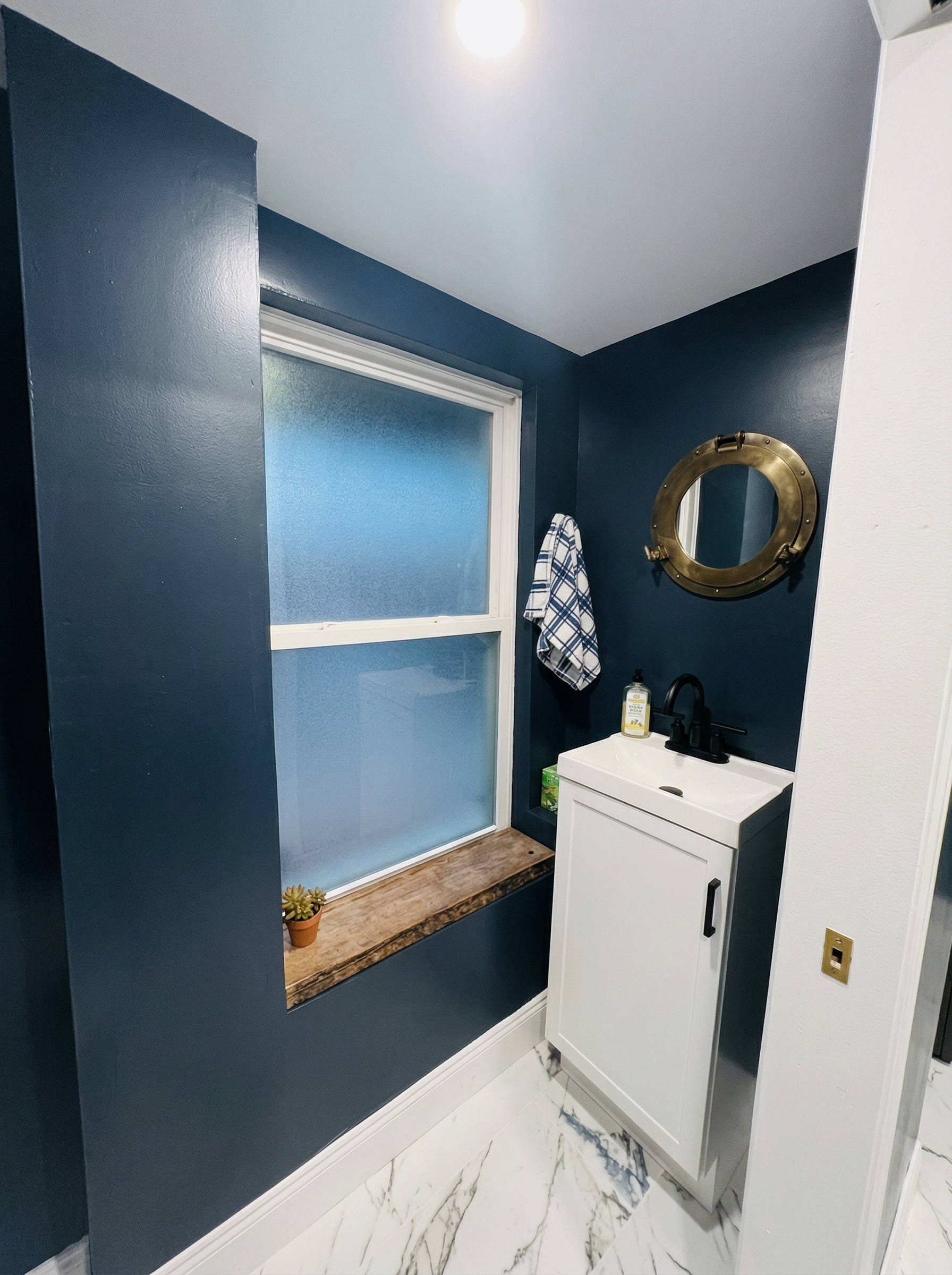A bathroom with dark blue walls, white trim, and a white vanity sink. The floor has white marble tiles with gray veining. There is a small potted succulent plant near a wooden window ledge, a towel hanging on a hook, and a gold porthole mirror on the